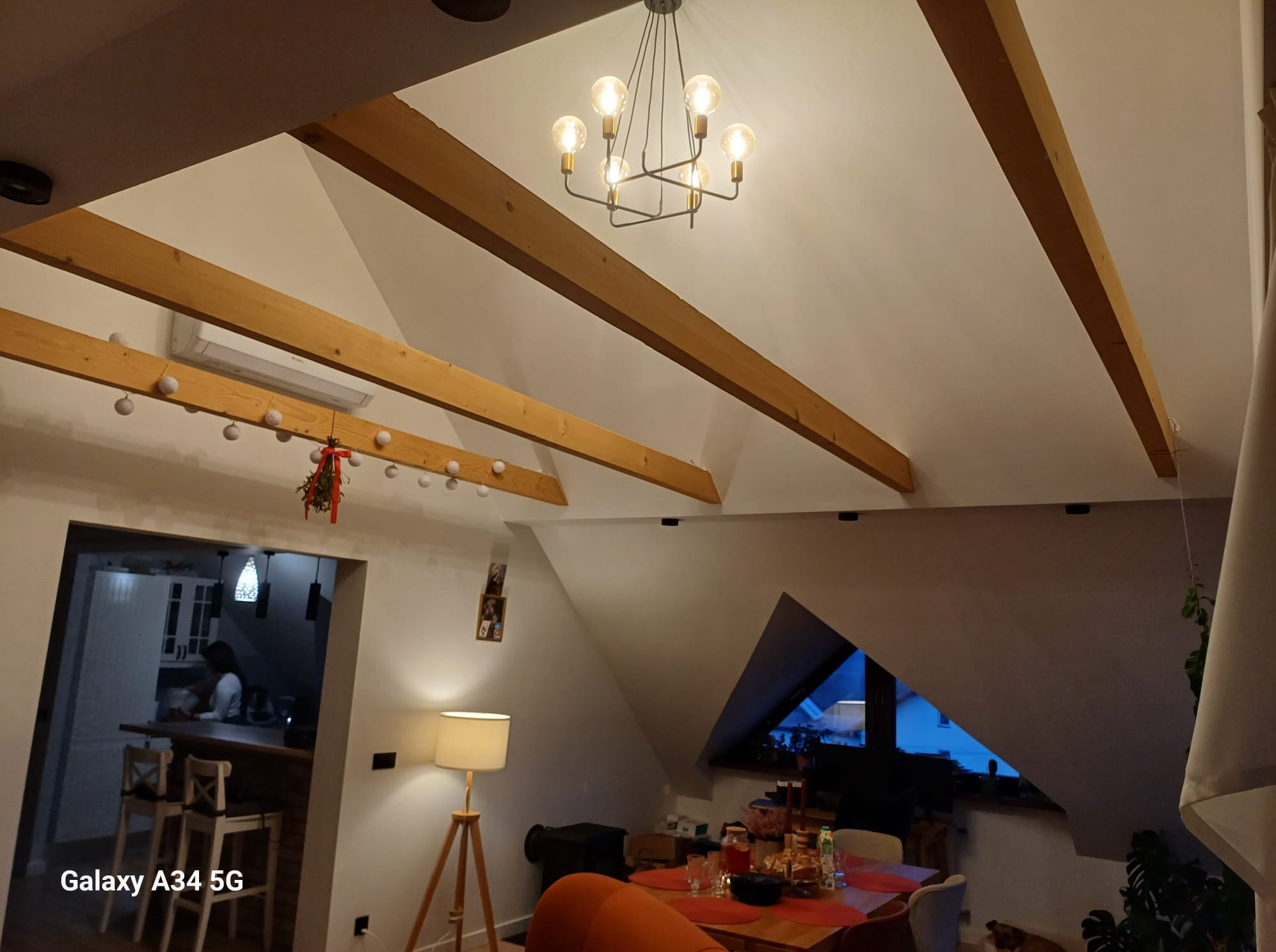 Jasne wnętrze poddasza z drewnianymi belkami stropowymi i nowoczesnym żyrandolem. Widoczny fragment kuchni w tle, oświetlony lampą stojącą.