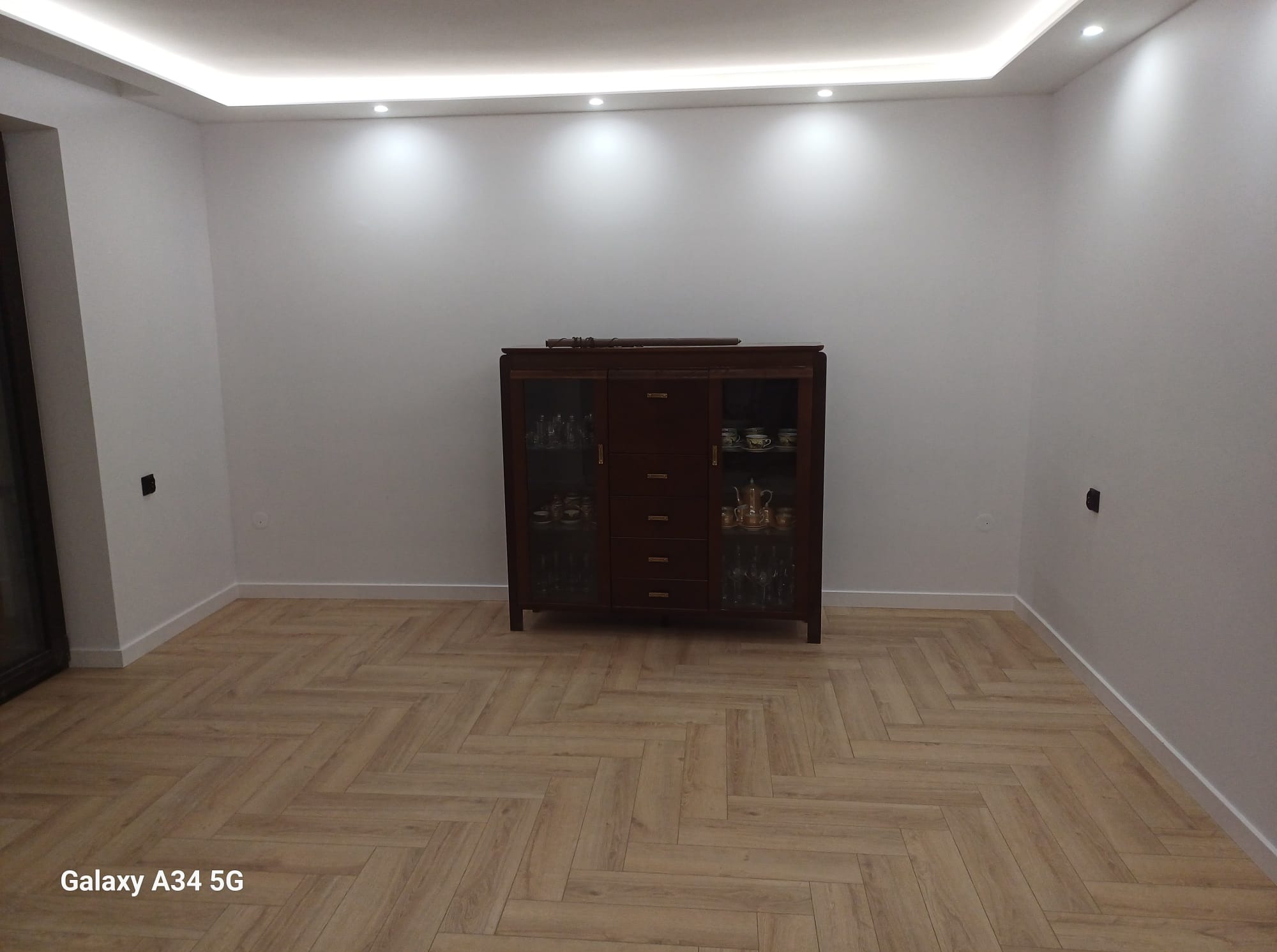 Pokój po remoncie: parkiet ułożony w jodełkę, białe ściany z listwami przypodłogowymi, oświetlenie sufitowe LED i ciemny kredens z przeszklonymi drzwiczkami.