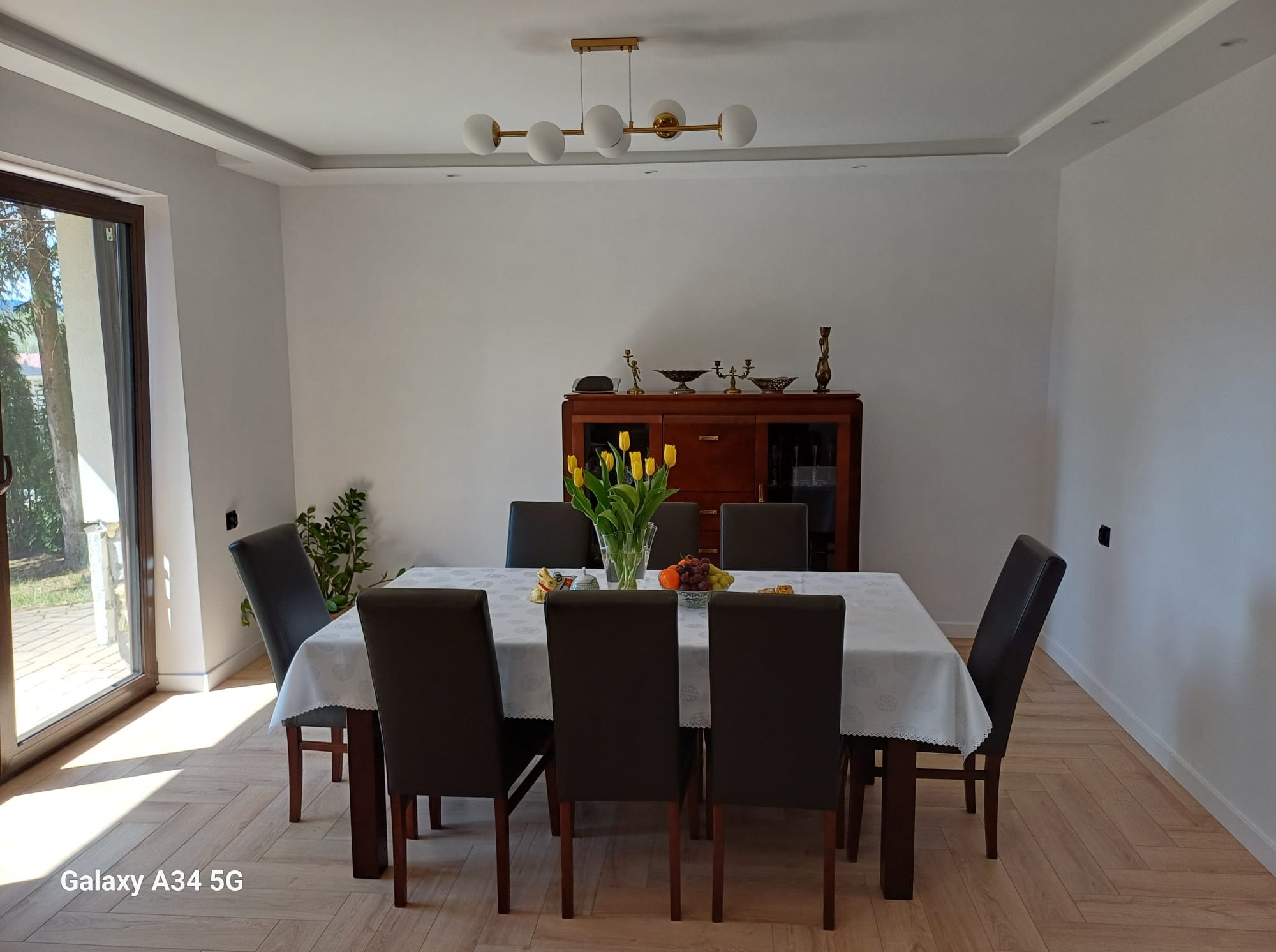 Jasne wnętrze jadalni z parkietem ułożonym w jodełkę, białymi ścianami, listwami przysufitowymi i złotym żyrandolem nad stołem z bukietem tulipanów.