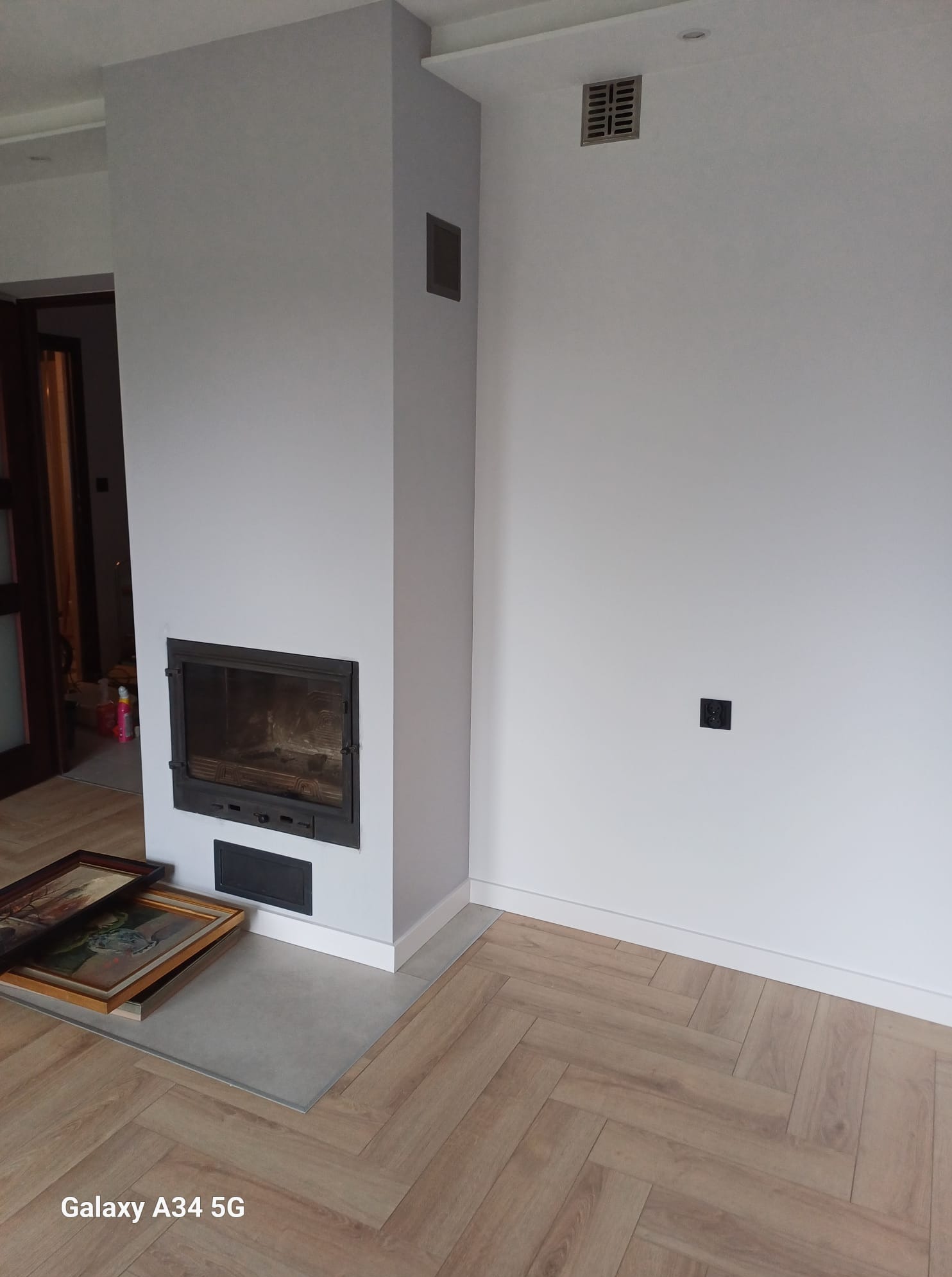 Minimalistyczny salon z kominkiem w obudowie, parkiet w jodełkę. Dwa obrazy w ramkach leżą na podłodze. Szara ściana, czarne gniazdko.