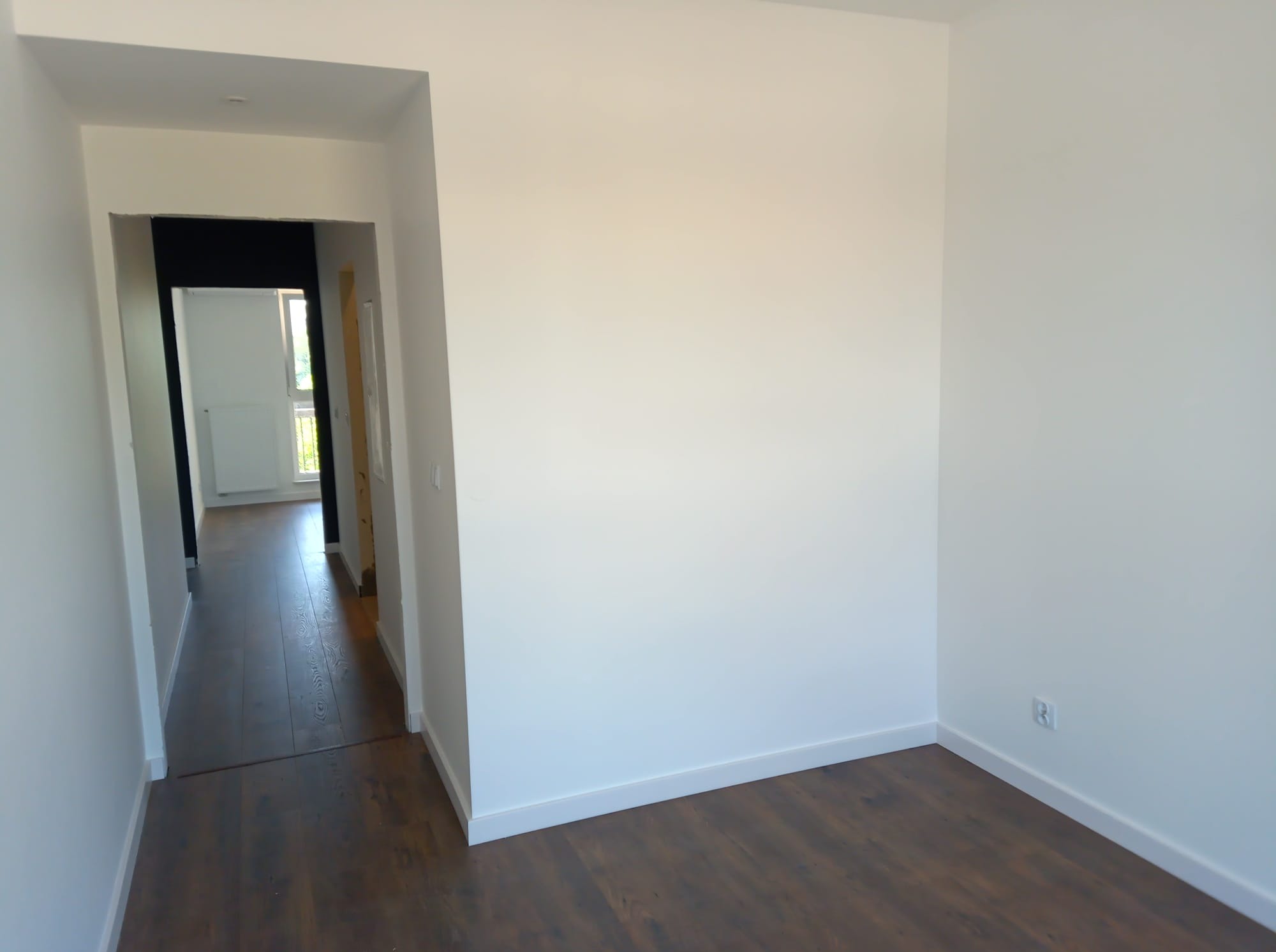 Pomalowane na biało wnętrze z ciemną podłogą, widoczne przejście do kolejnego pomieszczenia. Świeżo wykończone ściany i podłoga.