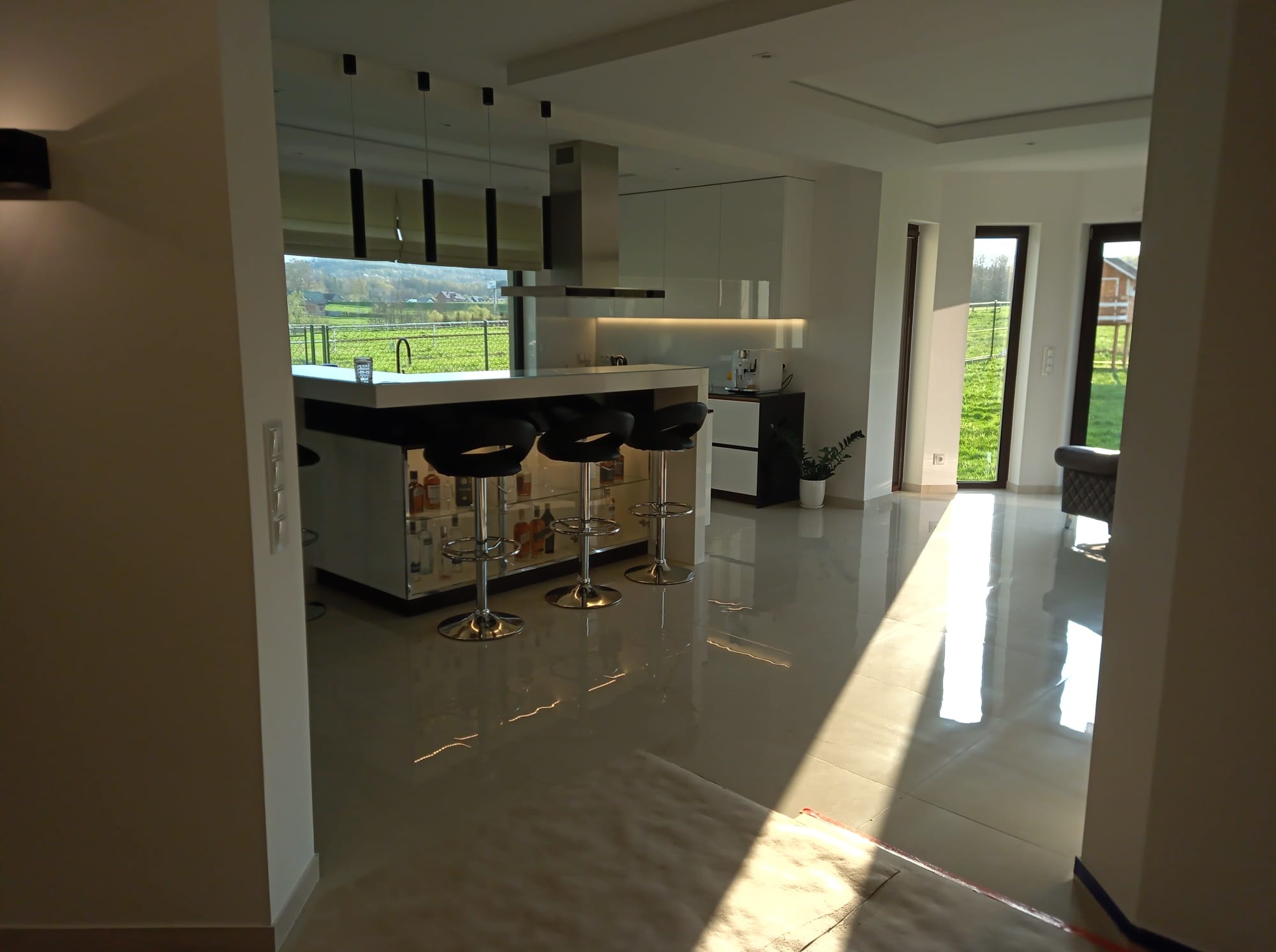 Nowoczesna kuchnia z wyspą i barkiem, widok z perspektywy wejścia. Błyszczące płytki podłogowe odbijają światło słoneczne. Minimalistyczne lampy wiszące nad blatem.