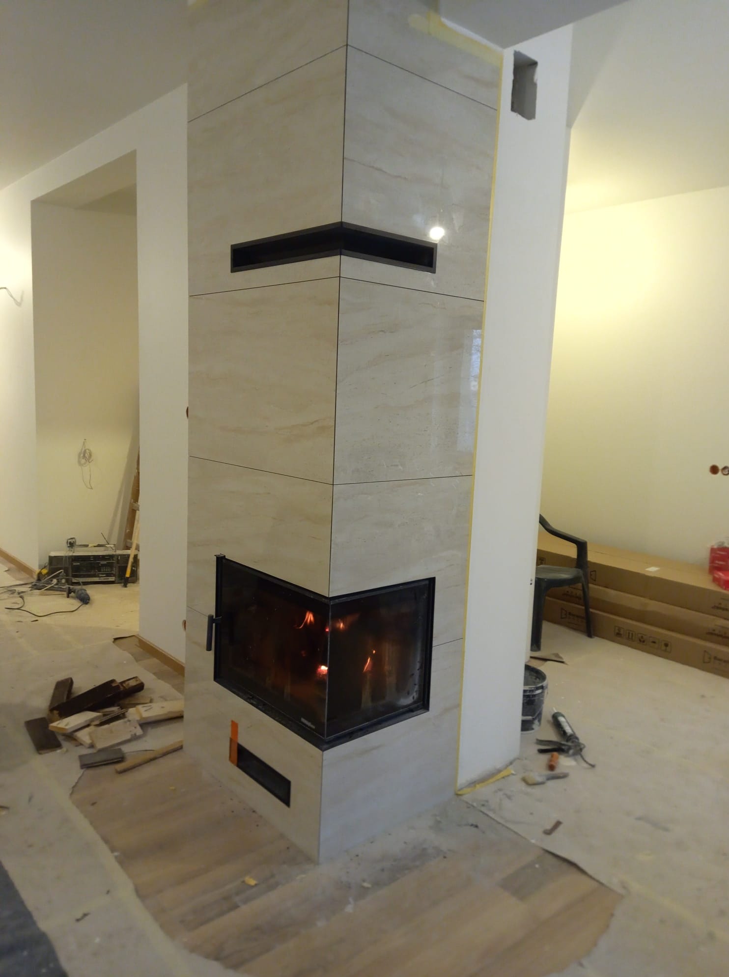 Obudowa kominka z beżowych, marmurkowych płyt w trakcie montażu. Widoczne narzędzia i materiały budowlane na podłodze. Otwarty ogień w palenisku.