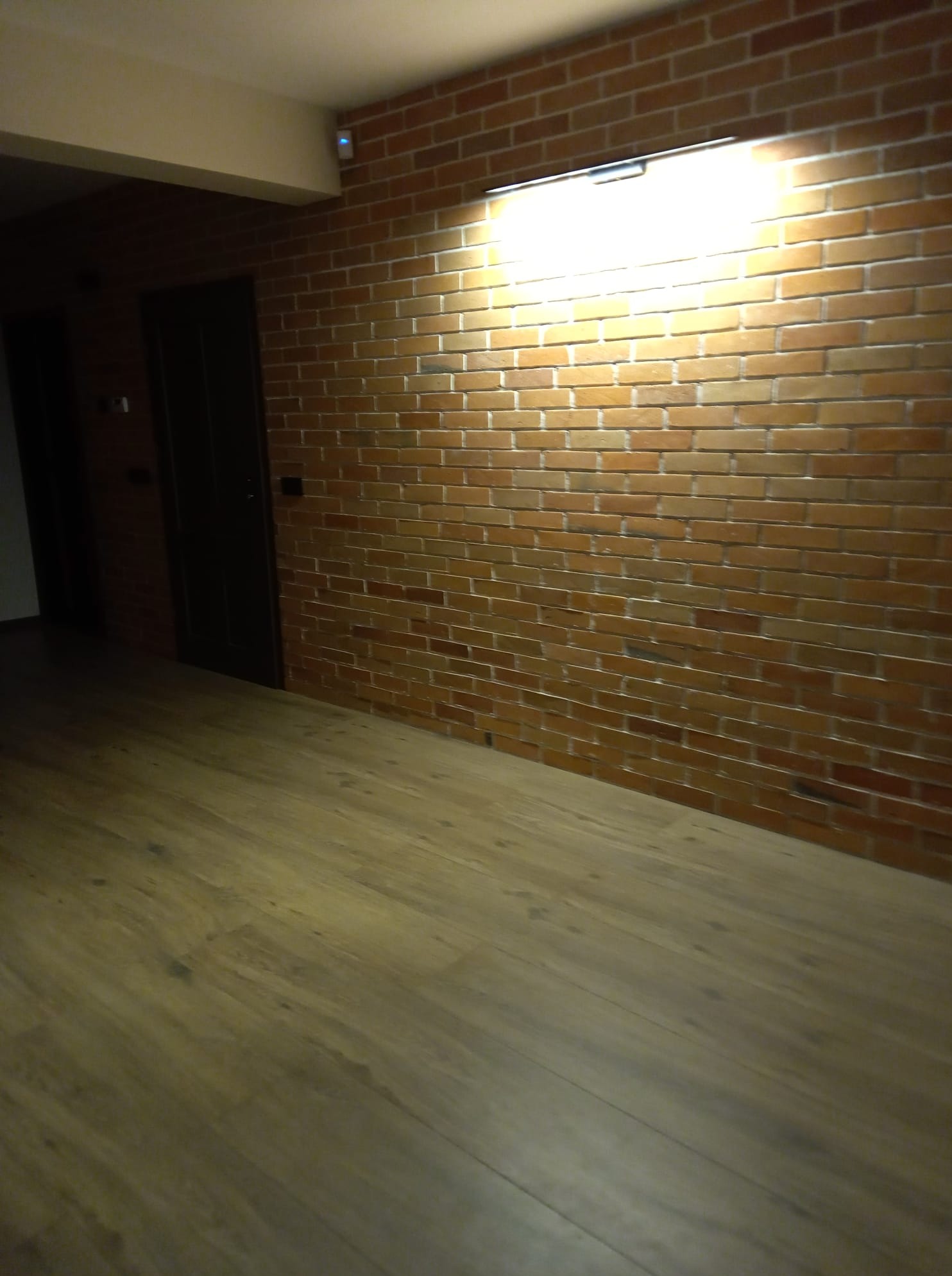 Ceglana ściana z oświetleniem i drewniana podłoga w Myślenicach. Widoczny fragment drzwi. Ciepłe oświetlenie nadaje wnętrzu przytulny charakter.