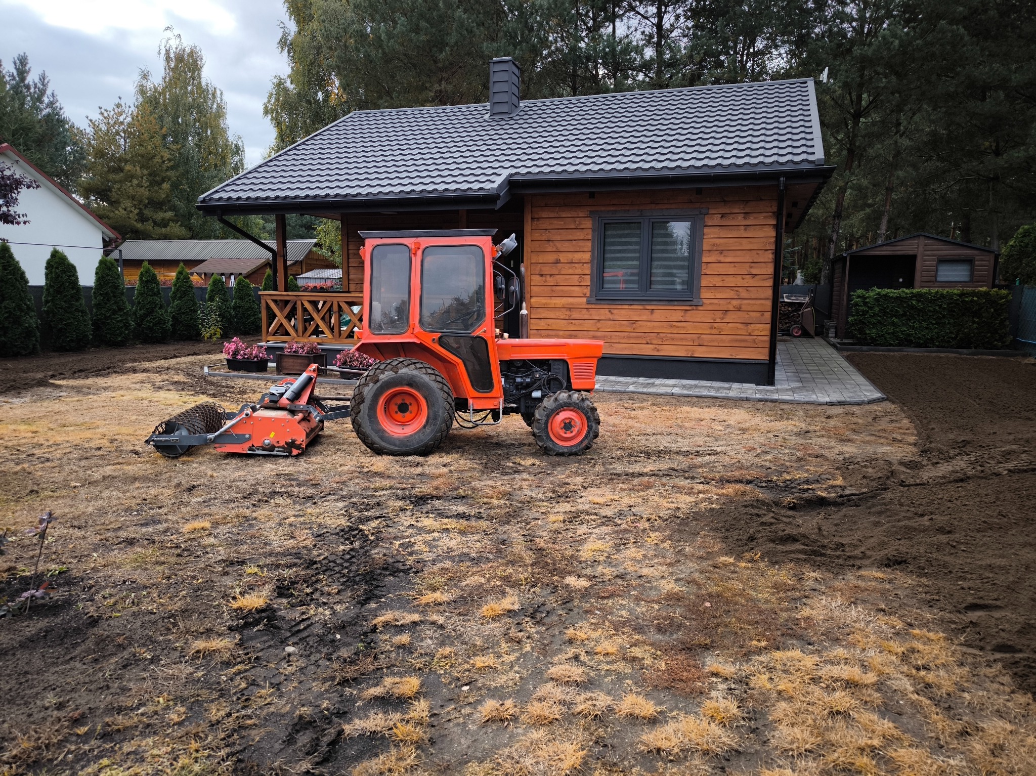 Czerwony traktor z wałem ugniatającym glebę na przygotowanym podłożu, obok drewniany dom z szarą dachówką i zagospodarowany teren.