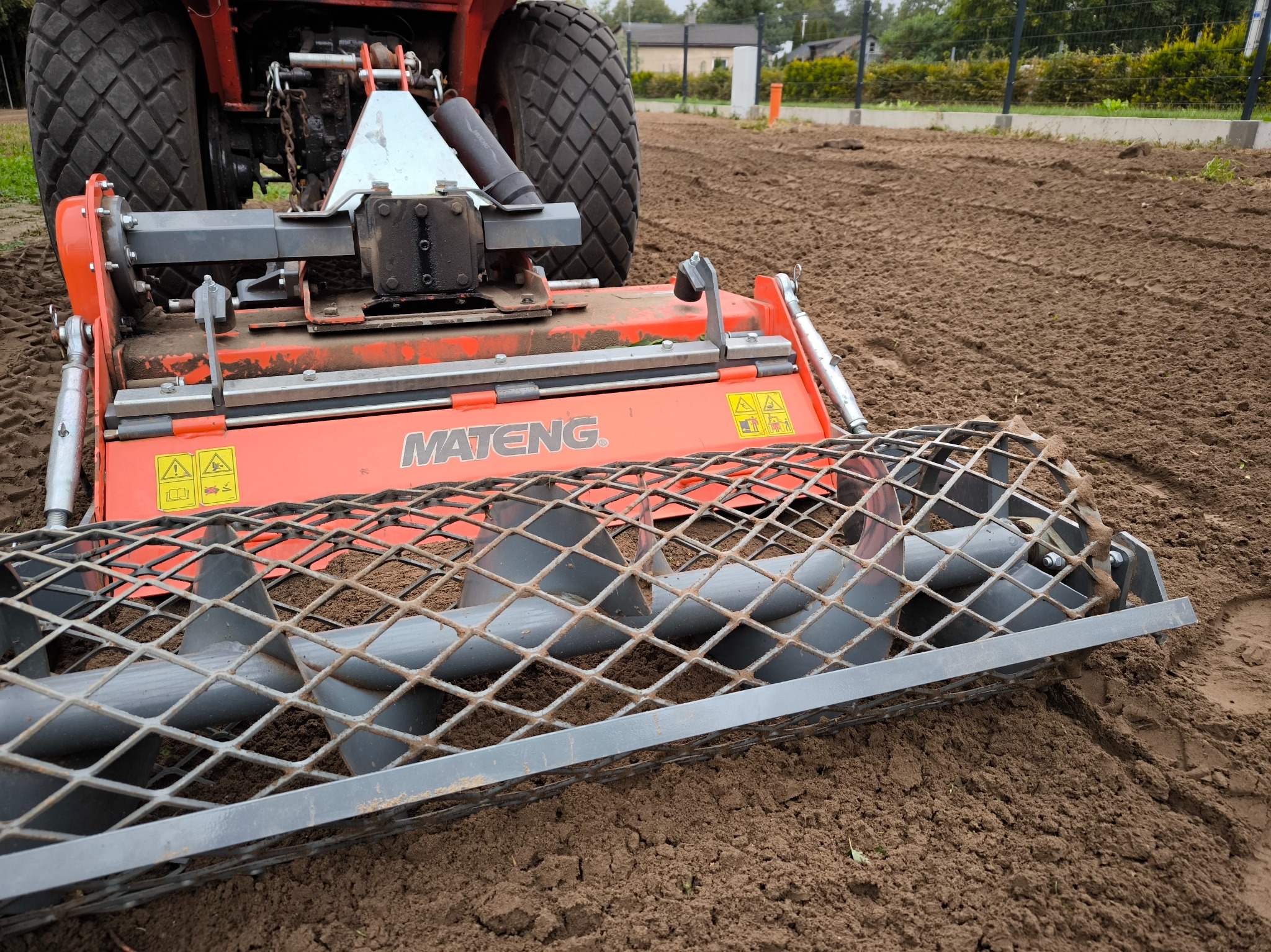 Czerwony traktor z glebogryzarką MATENG na przygotowanym polu, gotowym do siewu trawy. Widoczne ślady pracy urządzenia i wyrównana powierzchnia ziemi.
