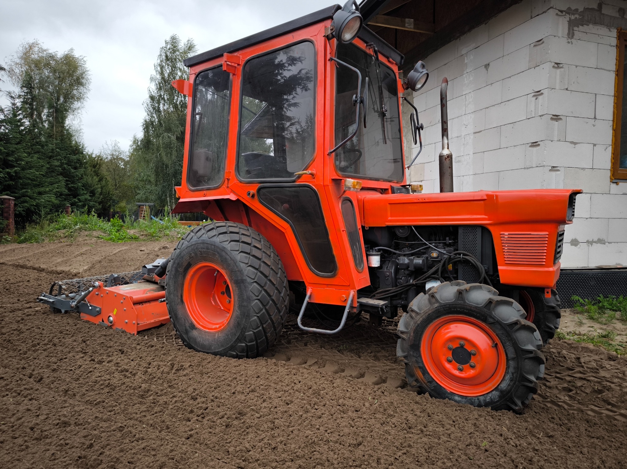 Czerwony traktor z glebogryzarką na przygotowanym polu, obok budynku z białych bloczków. Przygotowanie gruntu pod trawnik lub ogród.
