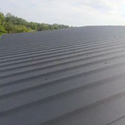 Szary dach z blachy trapezowej, widok z góry, z widocznymi śrubami mocującymi, na tle zieleni drzew i błękitnego nieba z lekkimi chmurami.