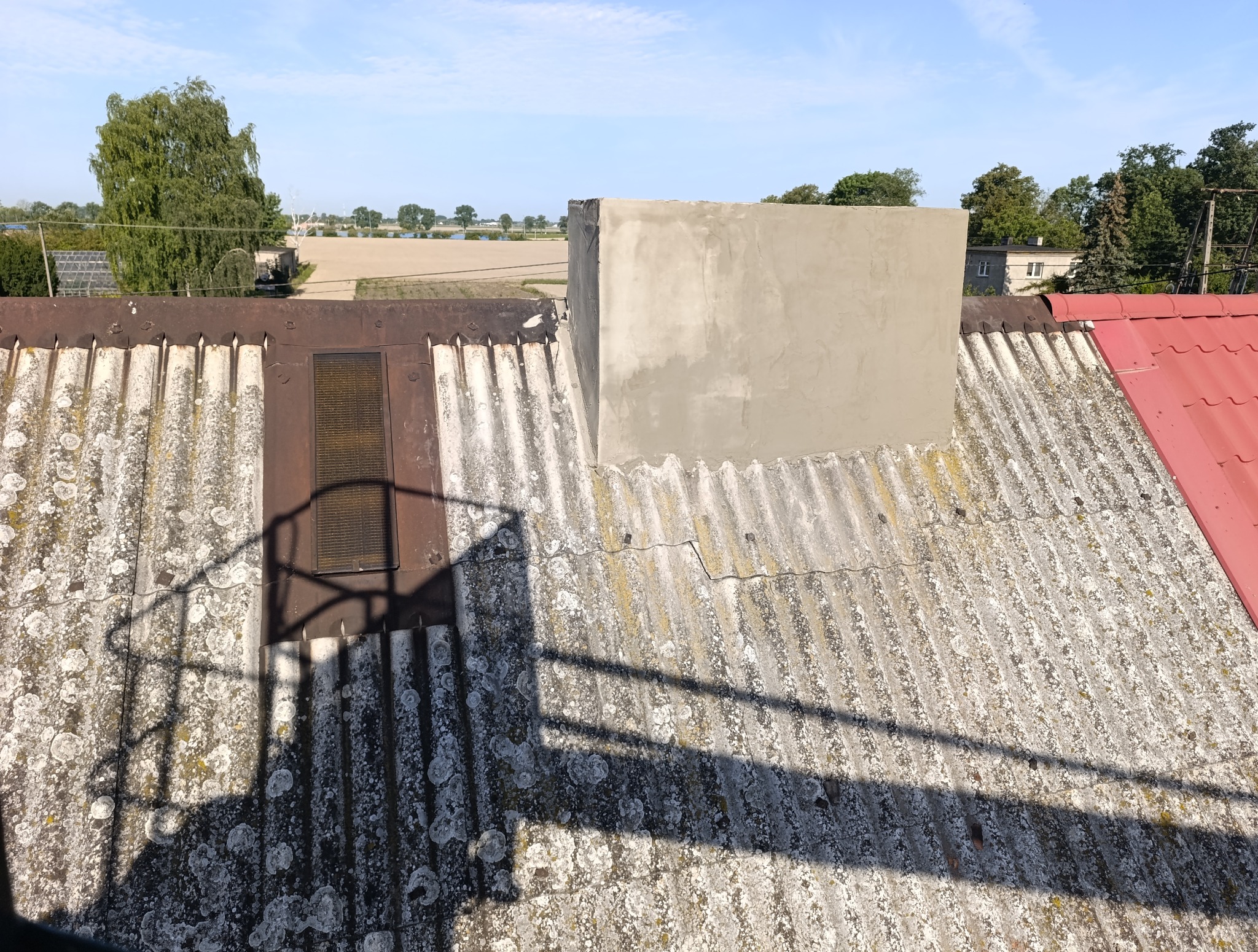 Szary dach z eternitu z kwadratową, otynkowaną konstrukcją kominową i fragmentem czerwonego dachu z blachy. Widoczne zacieki i porosty na eternicie. W tle drzewa i błękitne niebo.