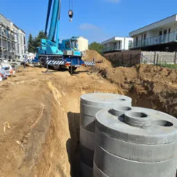 Montaż betonowych zbiorników w wykopie za pomocą dźwigu na placu budowy. Widoczne elementy konstrukcyjne budynków w tle i błękitne niebo.