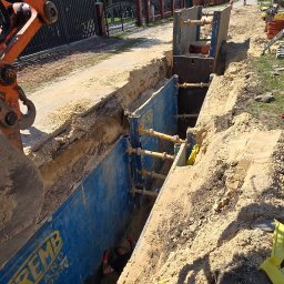 Garofano - Wykop zabezpieczony szalunkiem stalowym ZREMB, widoczny pracownik w wykopie. Na powierzchni pomarańczowe ramię koparki i elementy infrastruktury ulicznej.