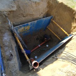 Garofano - Wykop zabezpieczony szalunkiem, wewnątrz pracownicy montują rury kanalizacyjne. Widoczne kable i drabina. Ujęcie z góry, oświetlenie naturalne.