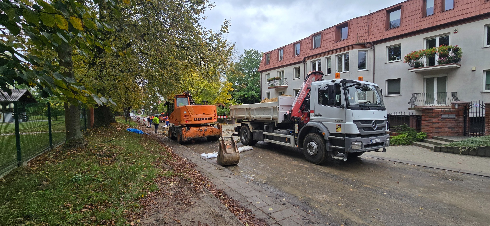 Pomarańczowa koparka Liebherr i ciężarówka z HDS na ulicy w Lublinie. Roboty ziemne w toku, widoczni pracownicy w odblaskowych kamizelkach. Jesienne drzewa w tle.
