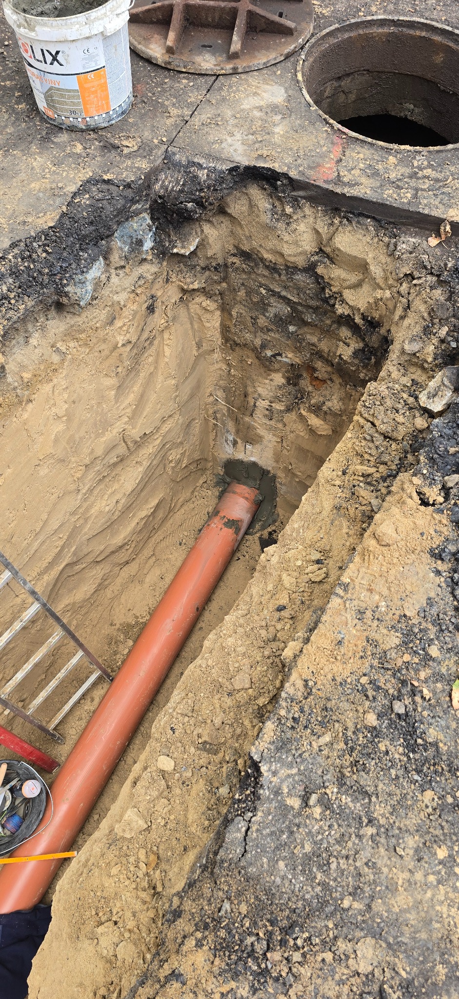 Wykop z rurą kanalizacyjną, częściowo zasypany, z widoczną drabiną i pokrywą studzienki. Na górze wiadro z zaprawą. Kąt ujęcia z góry, oświetlenie naturalne.