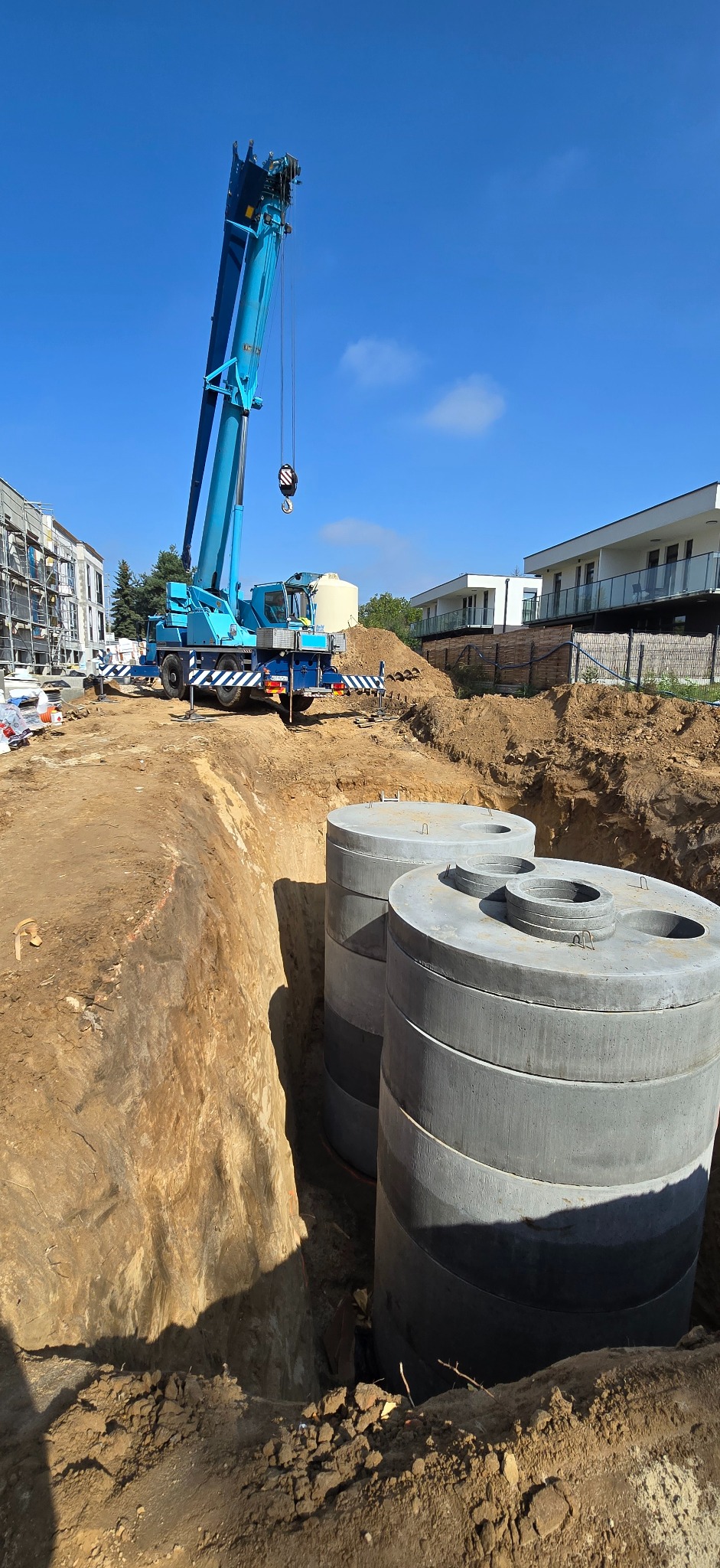 Montaż betonowych zbiorników w wykopie za pomocą dźwigu na placu budowy. Widoczne elementy konstrukcyjne budynków w tle i błękitne niebo.