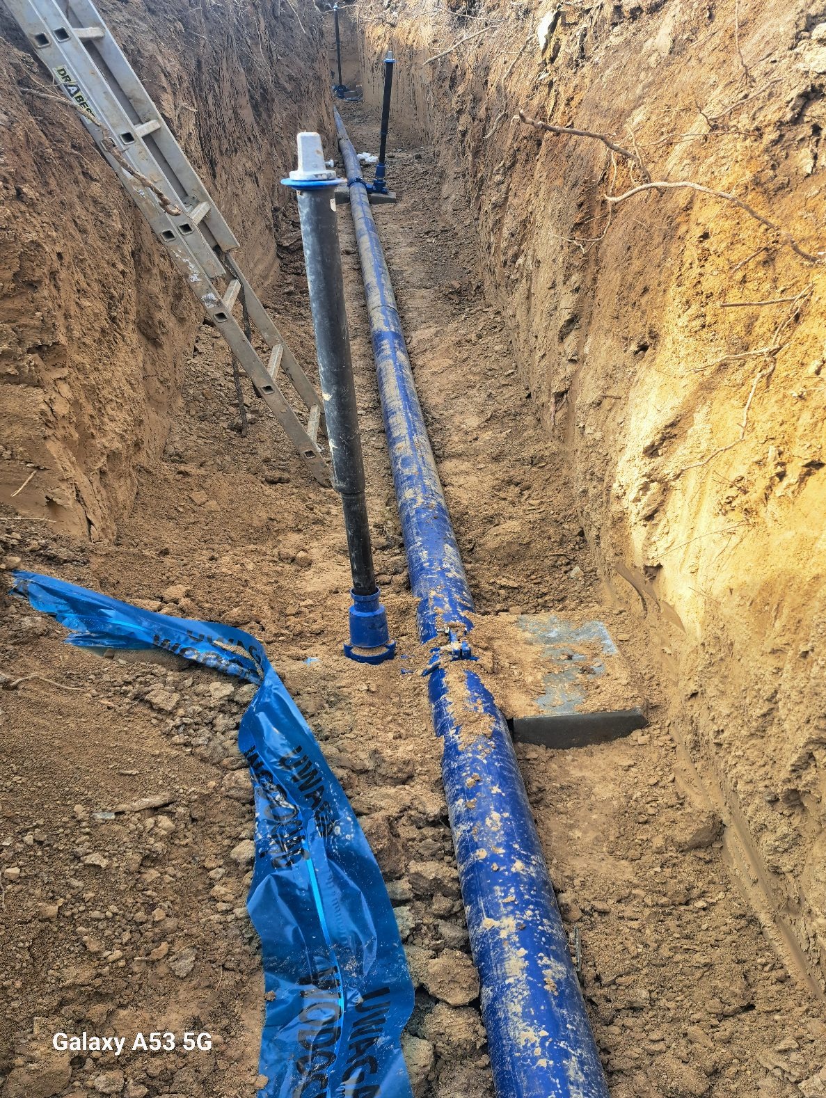 Wykop z ułożonymi niebieskimi rurami wodociągowymi, widoczna drabina i taśma ostrzegawcza. Grunt piaszczysty.