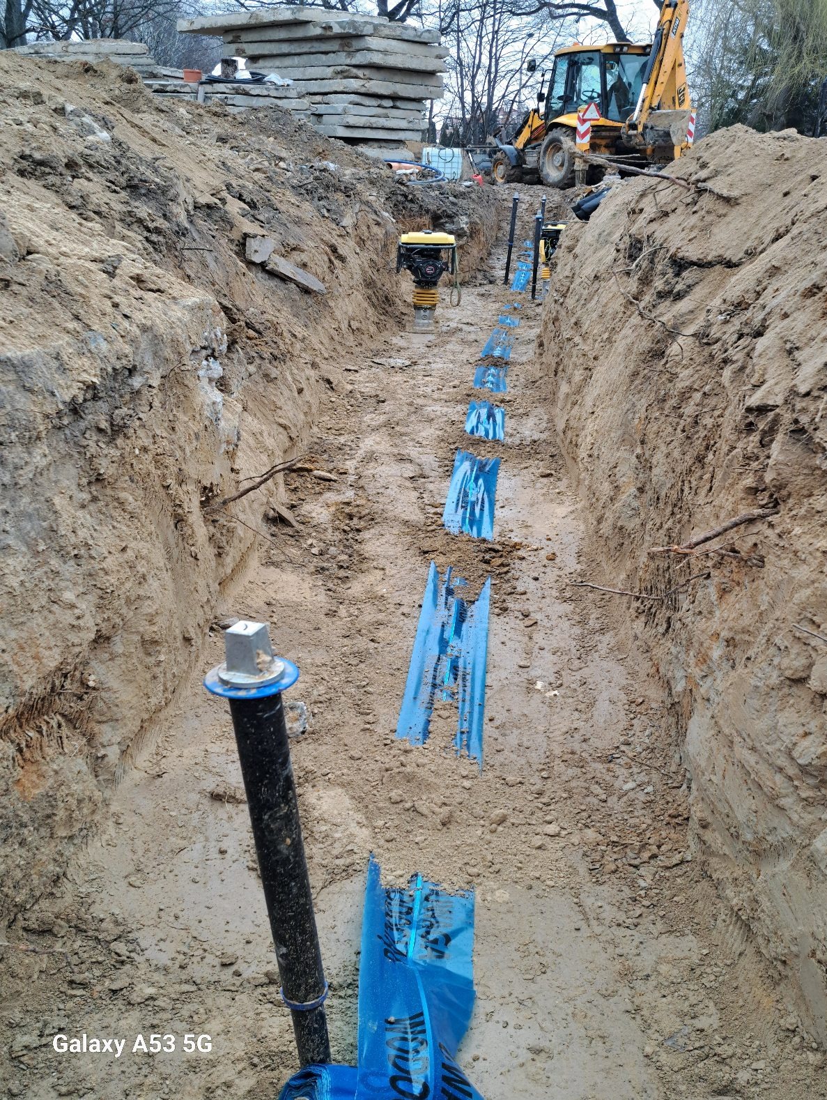 Wykop pod instalację wod-kan z widoczną rurą i niebieskimi elementami stabilizującymi. W tle zagęszczarka i koparka. Prace ziemne w trakcie realizacji.