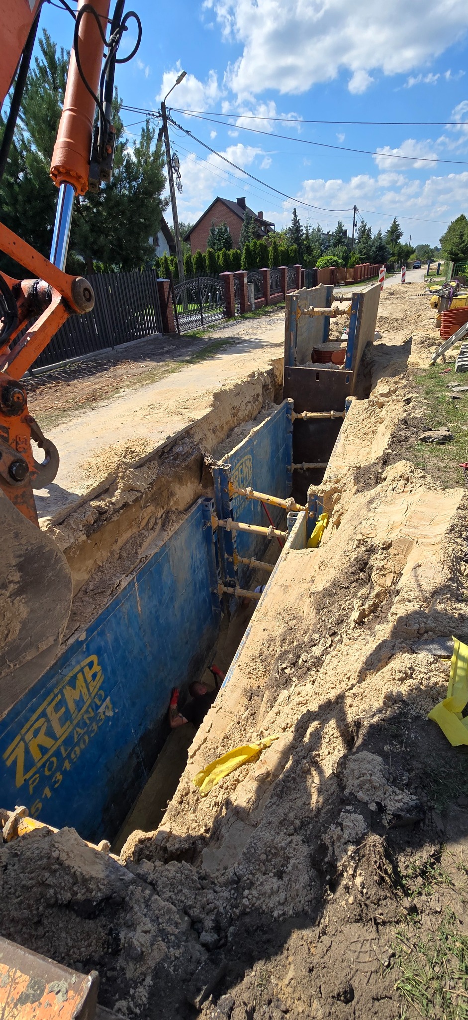 Wykop zabezpieczony szalunkiem stalowym ZREMB, widoczny pracownik w wykopie. Na powierzchni pomarańczowe ramię koparki i elementy infrastruktury ulicznej.