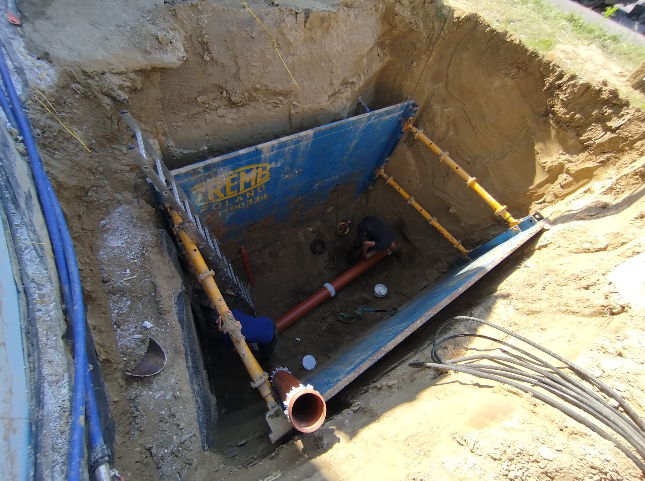 Wykop zabezpieczony szalunkiem, wewnątrz pracownicy montują rury kanalizacyjne. Widoczne kable i drabina. Ujęcie z góry, oświetlenie naturalne.