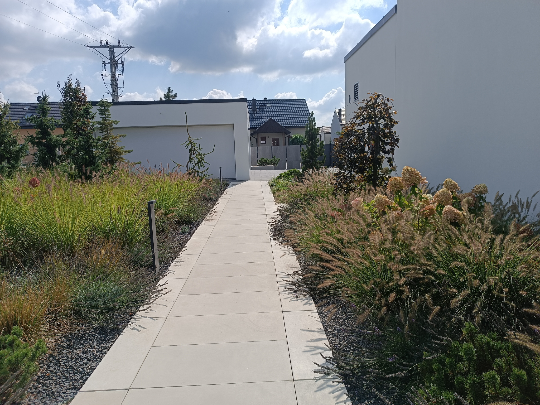Jasna, nowoczesna ścieżka z płyt, otoczona ozdobnymi trawami i krzewami, prowadzi do domu z garażem. Widoczne elementy architektury i słup energetyczny w tle.