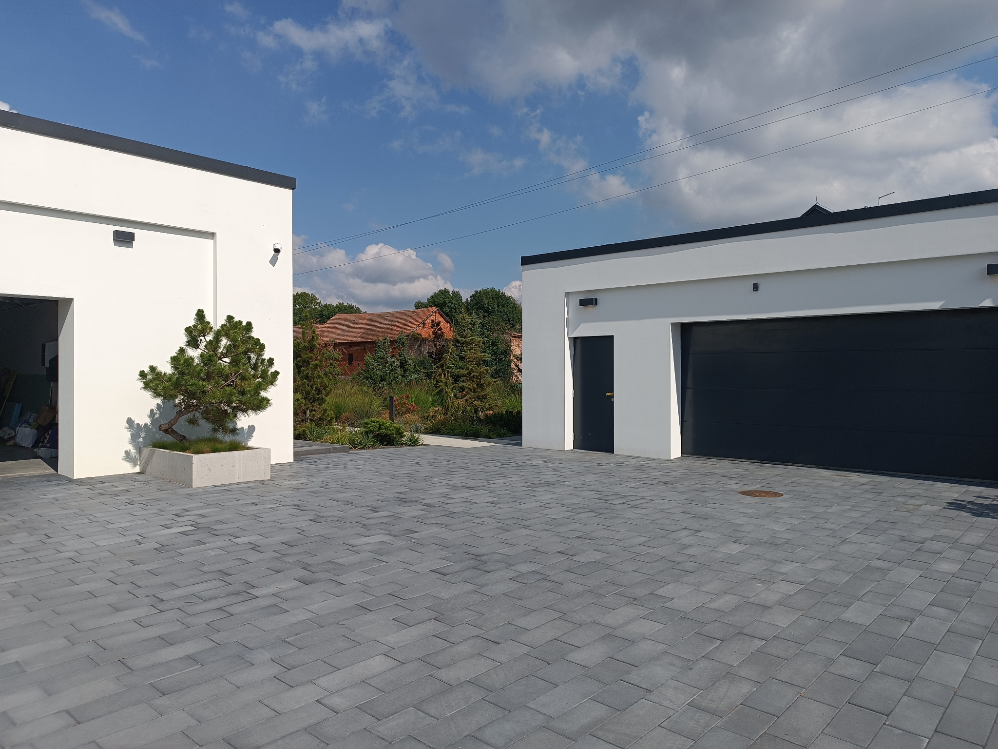 Szara kostka brukowa na podjeździe między dwoma nowoczesnymi budynkami z garażami w słoneczny dzień. W tle widoczny budynek z czerwoną dachówką.