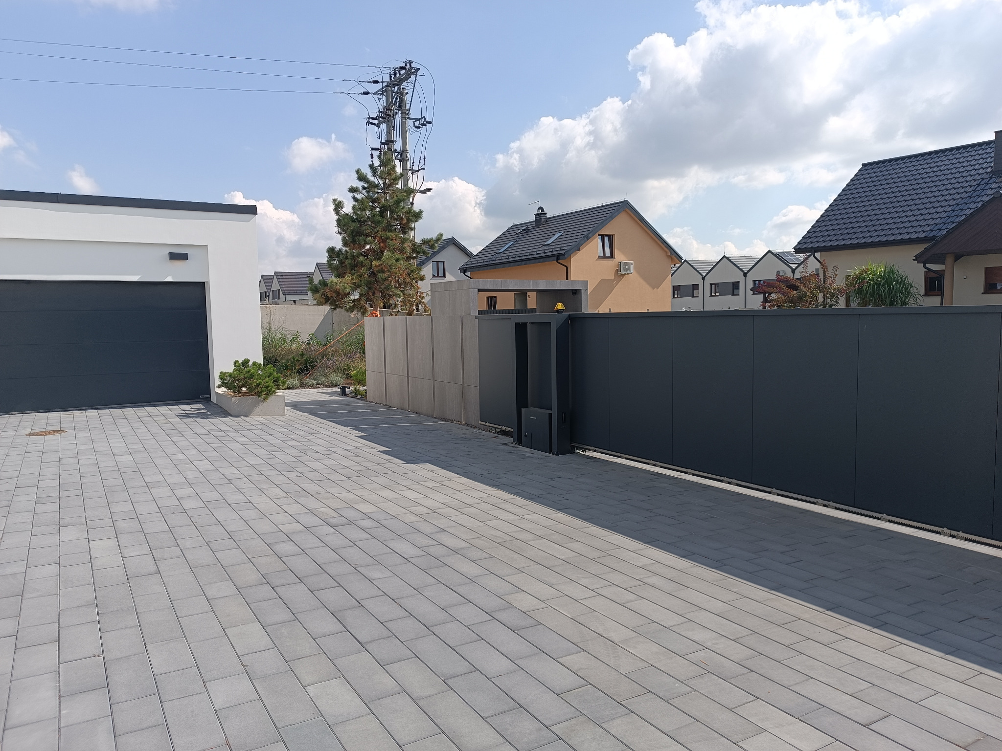 Nowoczesny podjazd z szarej kostki brukowej, brama przesuwna i garaż w tle. Widoczne domy jednorodzinne i słup energetyczny. Słoneczny dzień.