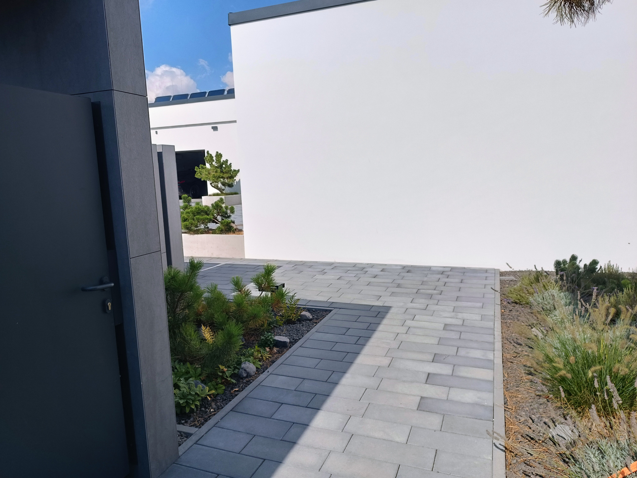 Nowoczesna posesja z szarą kostką brukową, minimalistycznym budynkiem i elementami zieleni, widok zza otwartych, grafitowych drzwi.