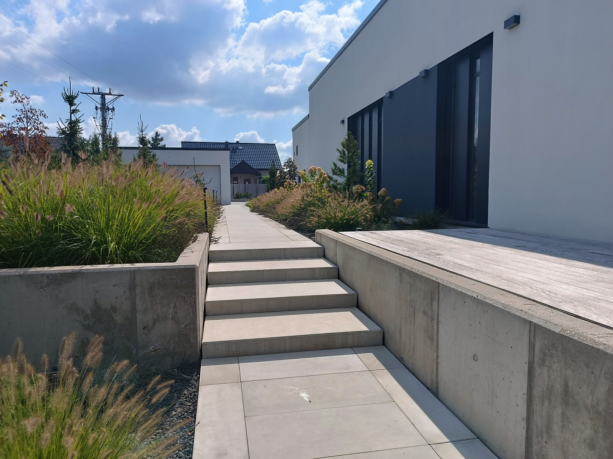 Nowoczesna ścieżka z jasnych płyt prowadzi przez ogród z ozdobnymi trawami do budynku z ciemnymi oknami. Minimalistyczny design i naturalne materiały.