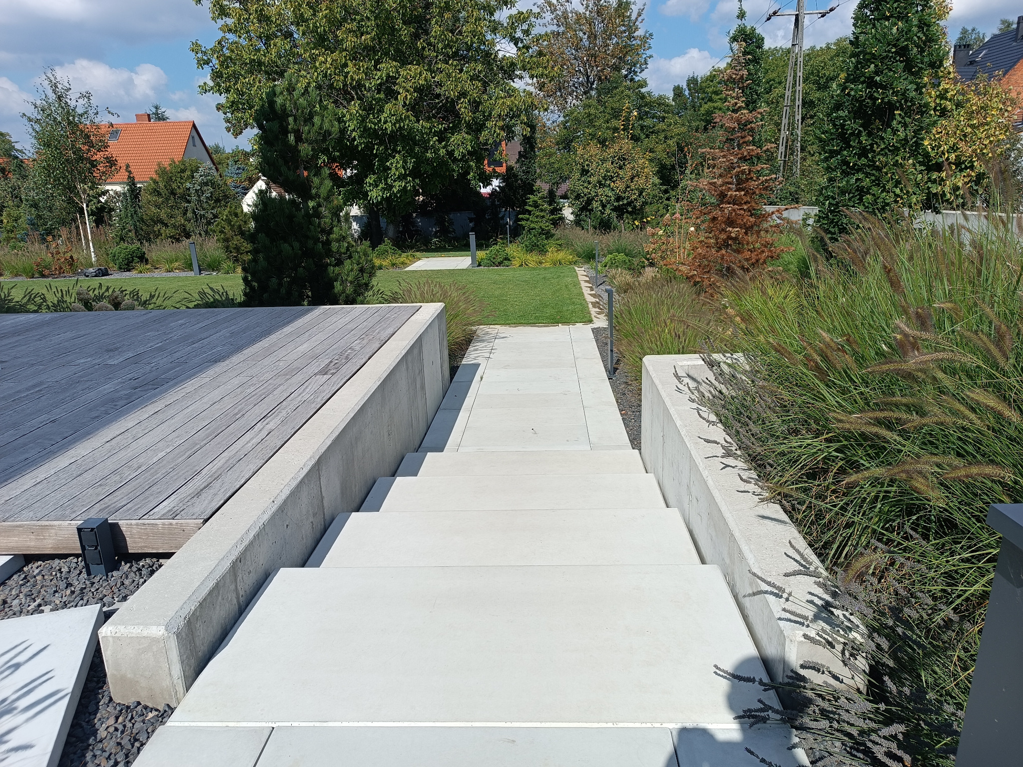 Nowoczesne schody z jasnych płyt betonowych prowadzą przez ogród z trawami ozdobnymi, łącząc drewniany taras z trawnikiem. Minimalistyczny design.