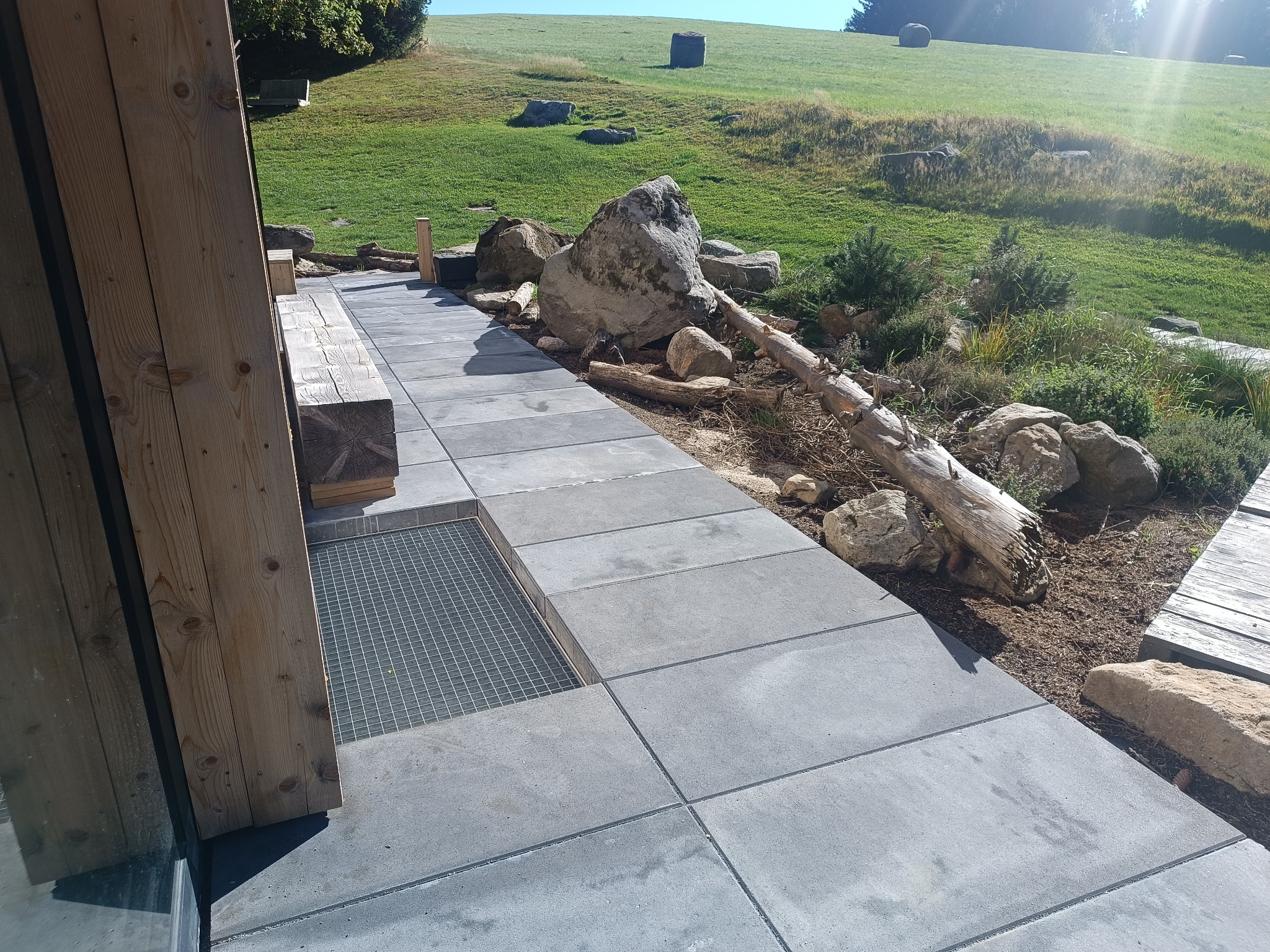 Nowoczesna ścieżka z szarej kostki brukowej prowadząca przez ogród, z elementami drewnianymi i kamieniami. Widoczna studzienka kanalizacyjna z kratą.