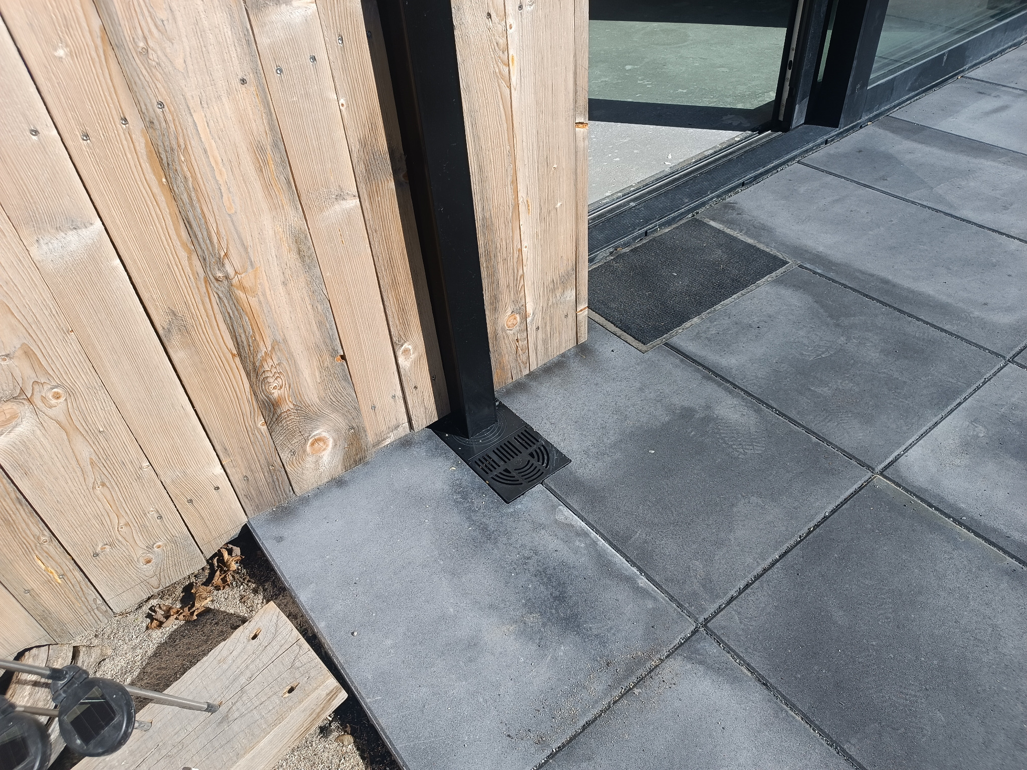 Szare płyty chodnikowe z odpływem liniowym przy drewnianym ogrodzeniu i progu drzwi przesuwnych. Widoczne liście i fragment lampy solarnej.