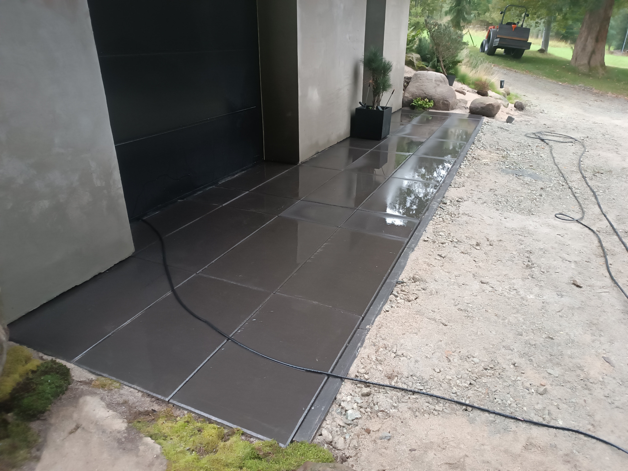 Nowoczesna, ciemna kostka brukowa ułożona przy budynku. W tle widać walec drogowy i teren przygotowany pod dalsze prace brukarskie. Minimalistyczny design.