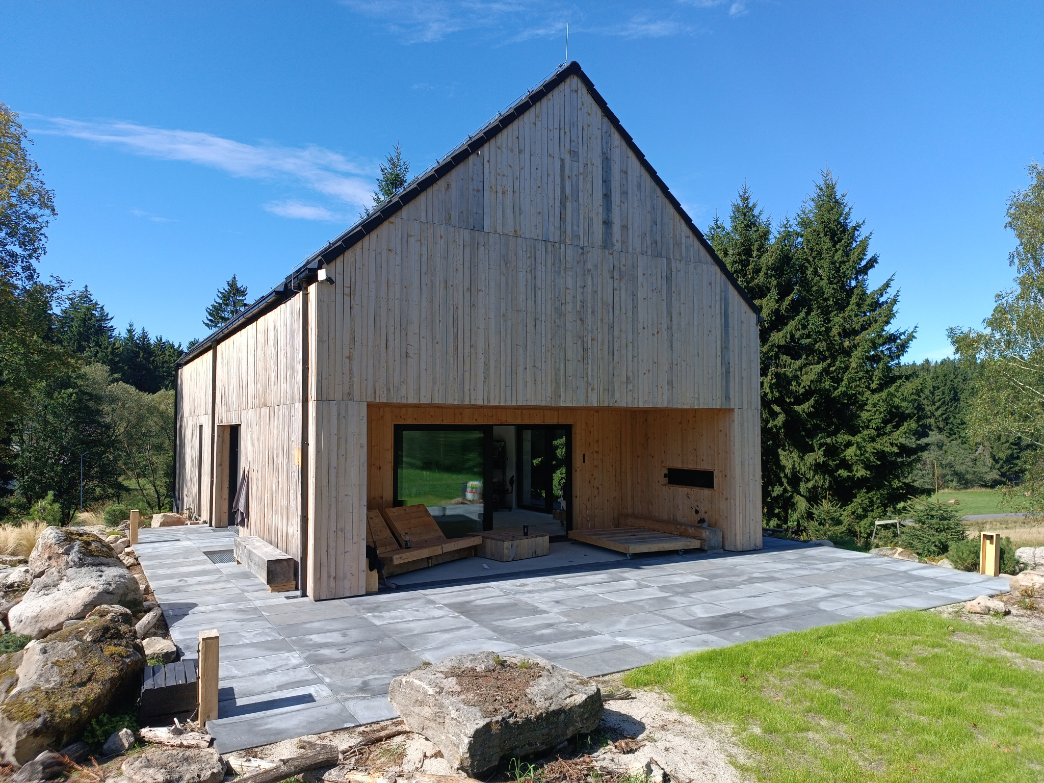 Nowoczesny dom z jasnego drewna z tarasem wyłożonym szarą kostką brukową. W tle zieleń drzew i błękitne niebo. Elementy krajobrazu wkomponowane w projekt.