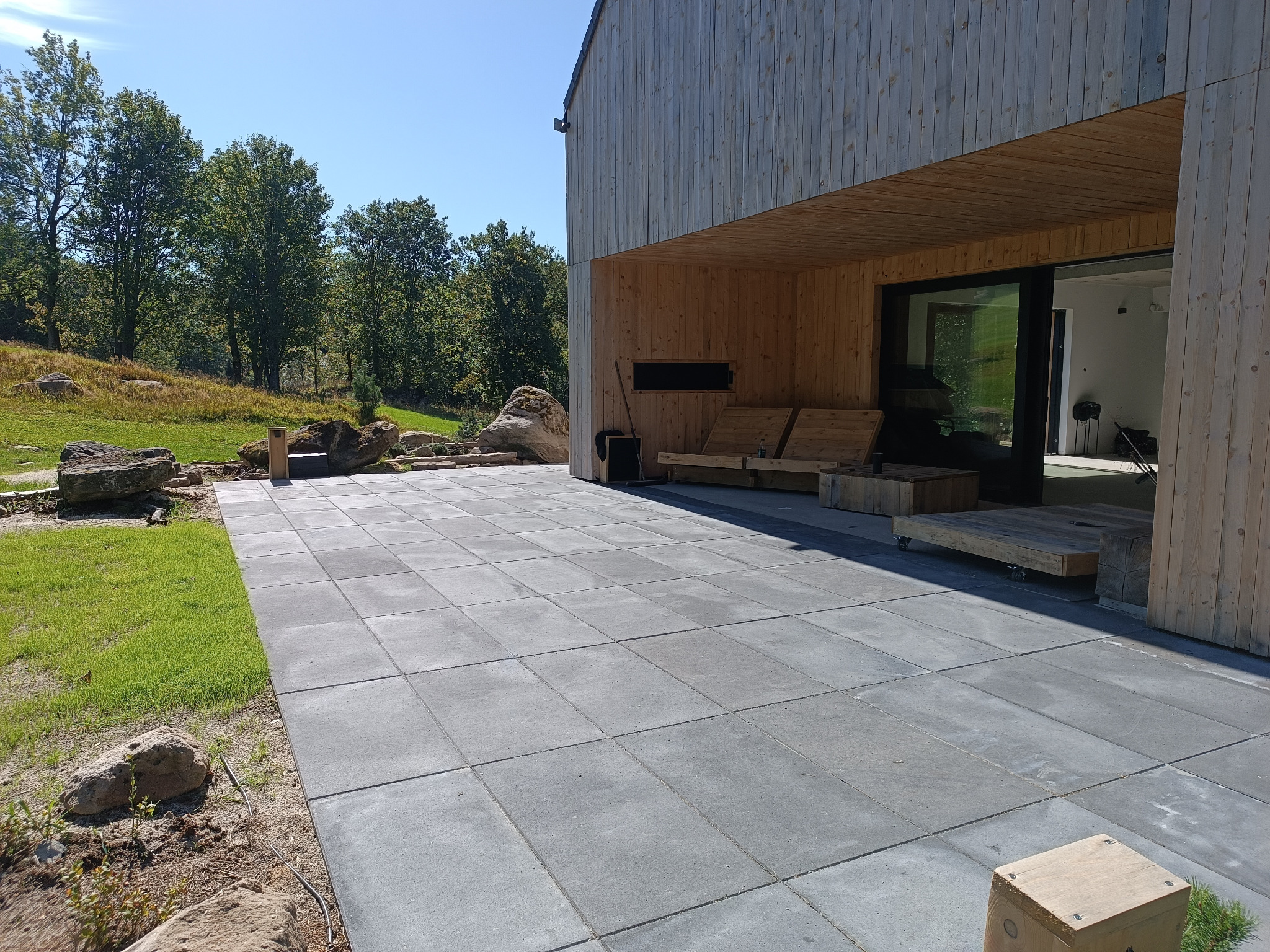 Nowoczesny taras z szarej kostki brukowej przy drewnianym domu, z elementami naturalnego krajobrazu: skały, trawa. Widok na las w tle, w słoneczny dzień.