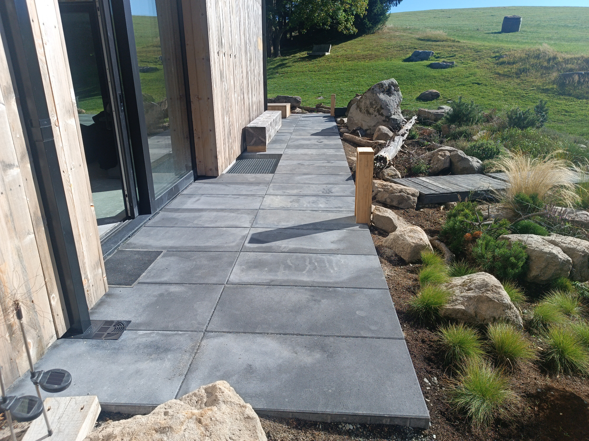 Nowoczesny taras z szarych płyt betonowych, harmonijnie wkomponowany w naturalny krajobraz z kamieniami i roślinnością. Widok na zielone wzgórze w tle.