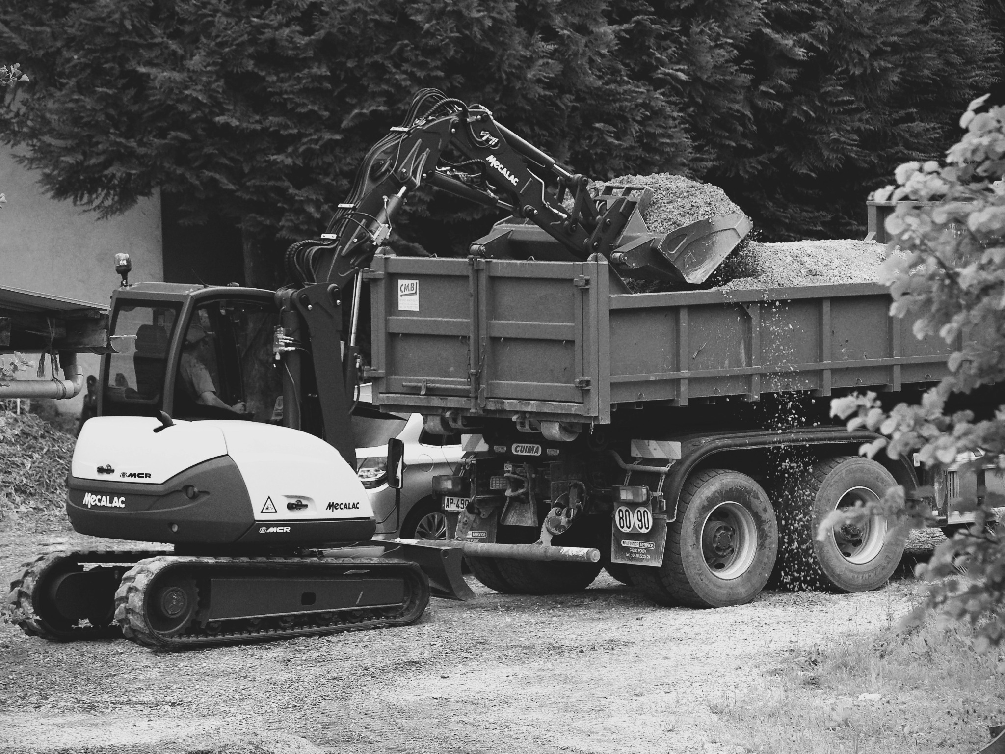 Czarno-białe zdjęcie: koparka Mecalac ładuje gruz do ciężarówki. Widoczne drzewa w tle. Praca wre, a gruz opada z łyżki koparki.