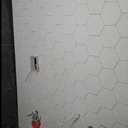 Dariusz Piwoński - Ściana z białych, heksagonalnych płytek w trakcie montażu, z widocznymi przewodami i elementami instalacji, na tle ciemnej, marmurowej ściany i surowej posadzki.