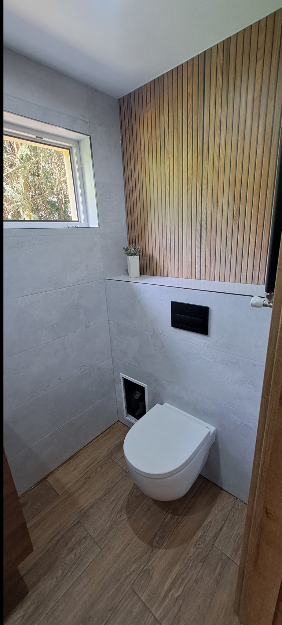 Nowoczesna łazienka z drewnianą ścianą, płytkami imitującymi kamień i podwieszaną toaletą. Okno z widokiem na zieleń. Mała roślina w białej doniczce.