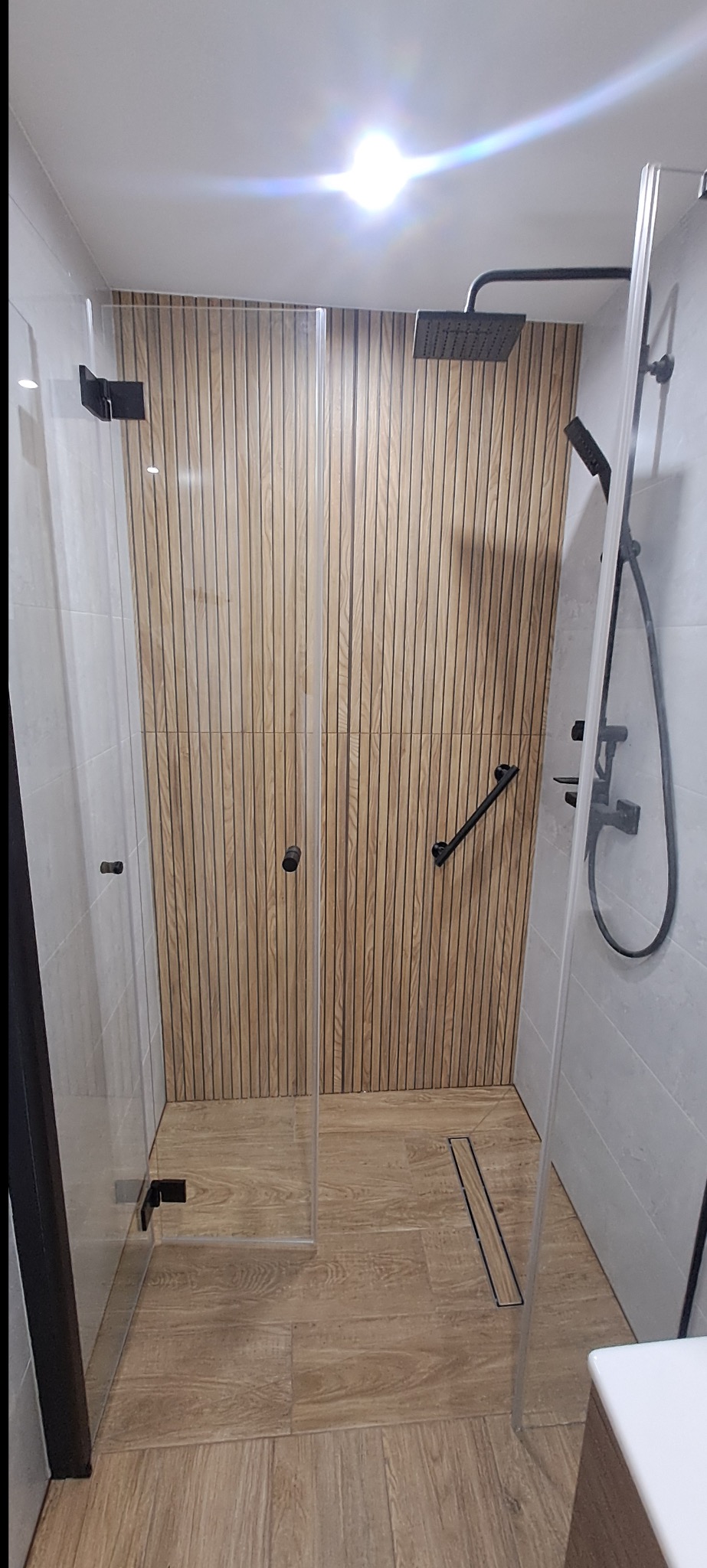 Nowoczesna kabina prysznicowa z drewnianym panelem na ścianie i czarną armaturą. Minimalistyczny design, jasne płytki i podłoga imitująca drewno.