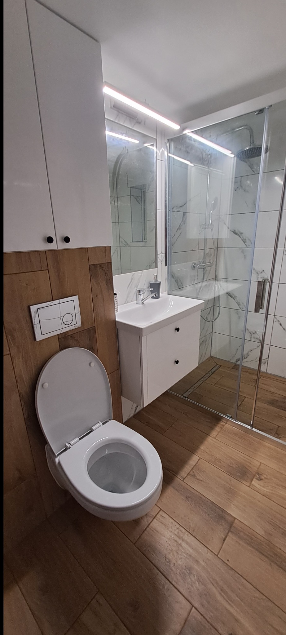 Nowoczesna łazienka z drewnopodobną podłogą, białą armaturą i kabiną prysznicową typu walk-in. Widoczna umywalka z szafką i toaleta z podtynkowym systemem spłukiwania.