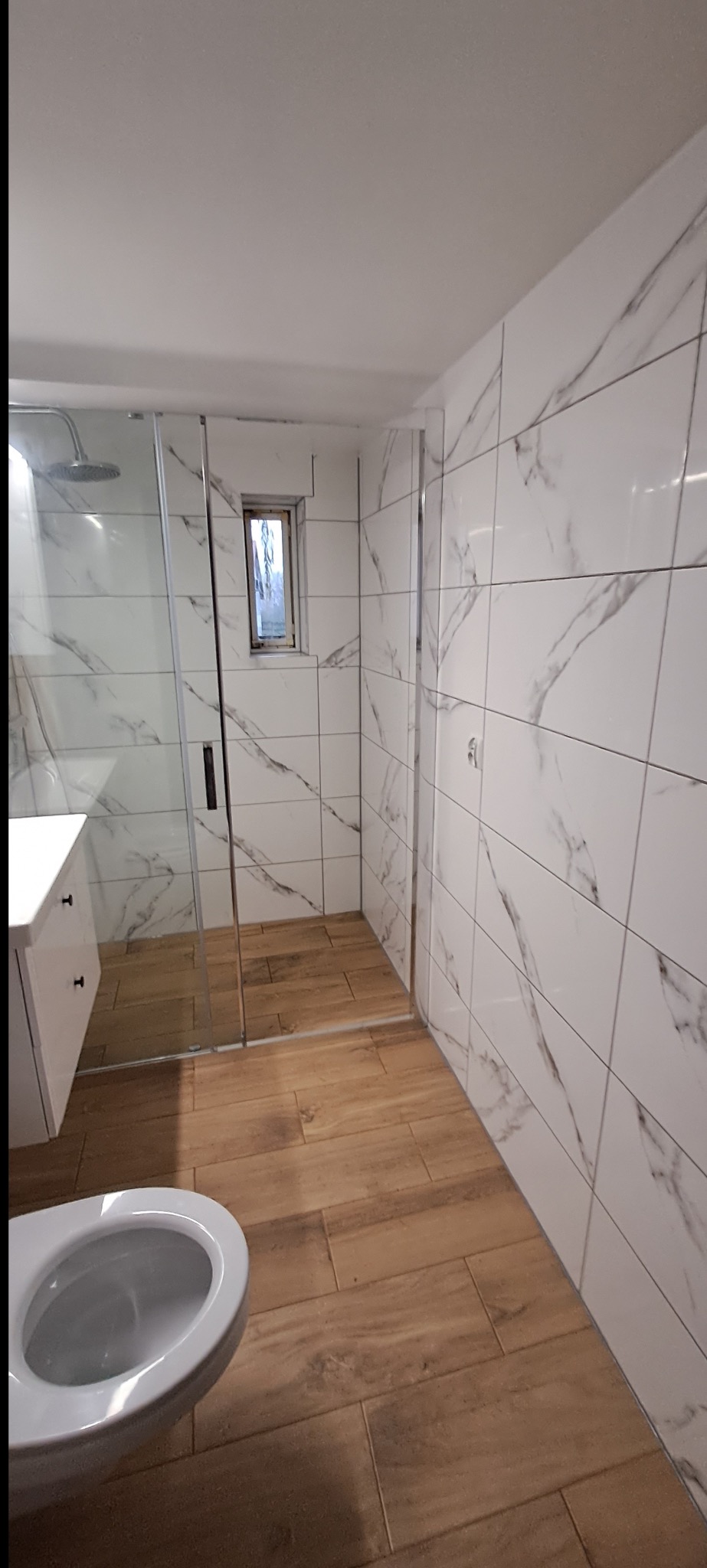 Nowoczesna łazienka po remoncie: kabina prysznicowa ze szklanymi drzwiami, białe płytki marmurowe na ścianach, drewnopodobna podłoga, toaleta i umywalka.