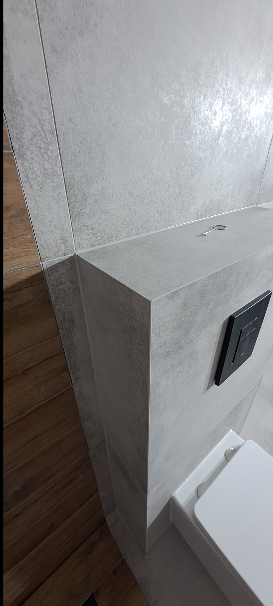 Wykończona toaleta podwieszana z szarą, błyszczącą płytką imitującą beton i drewnianą podłogą. Na obudowie toalety leży klucz.