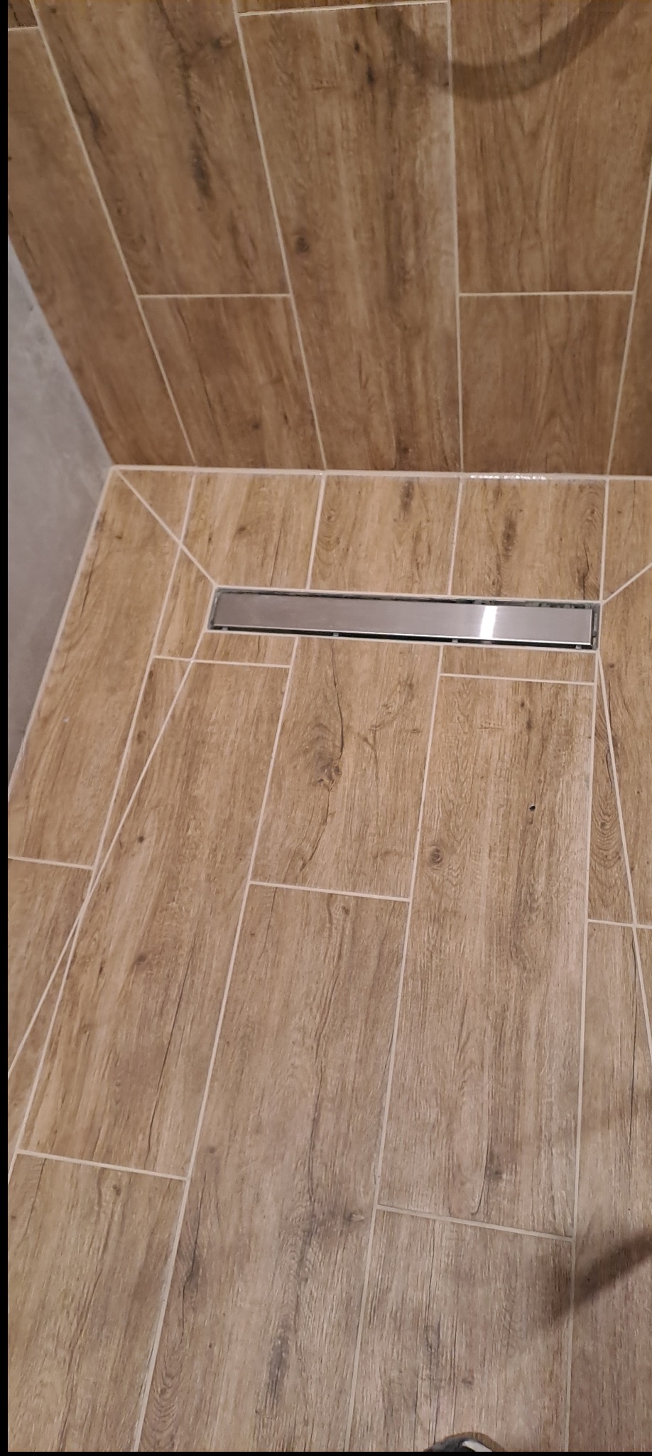 Brodzik z odpływem liniowym, wyłożony płytkami imitującymi drewno, ułożonymi w prostokątnym wzorze. Widok z góry, z bliska.