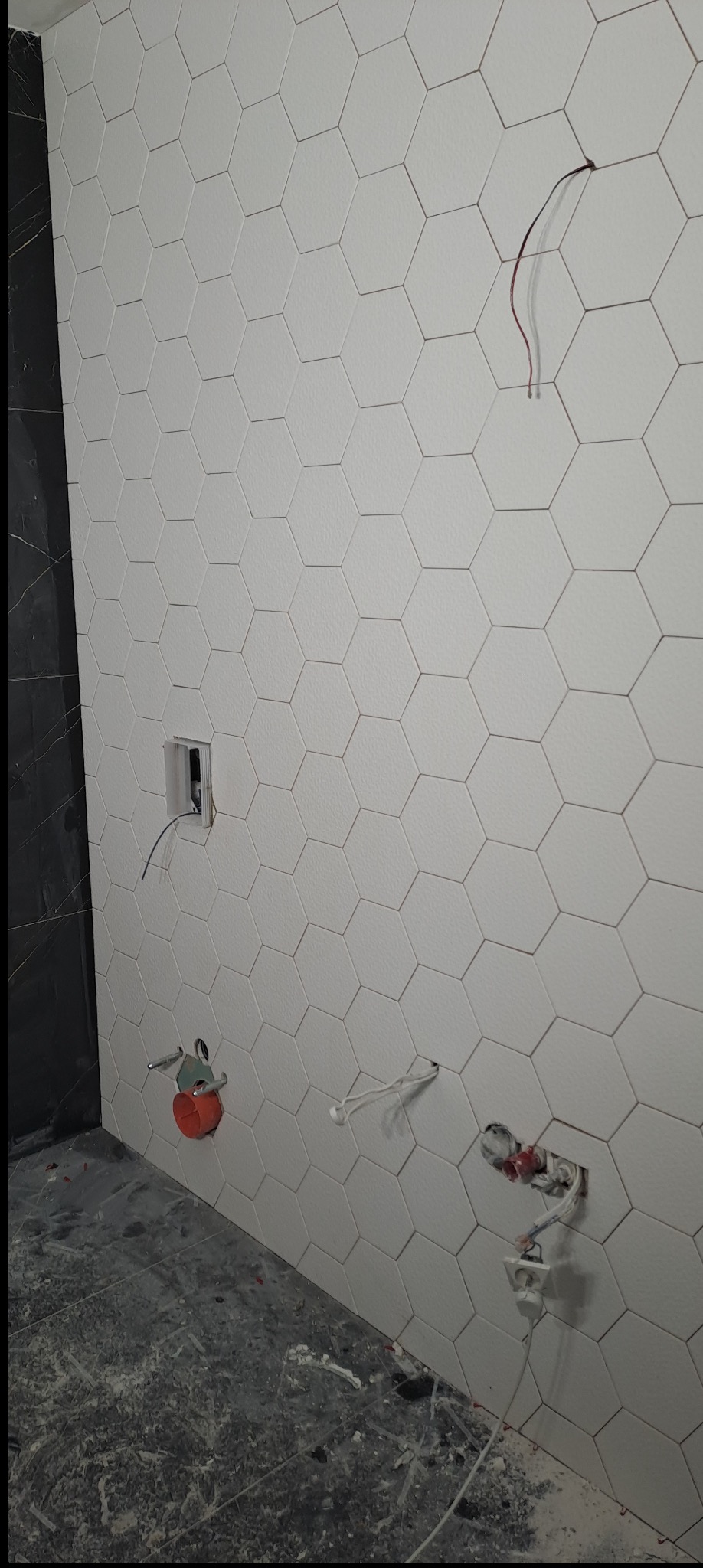 Ściana z białych, heksagonalnych płytek w trakcie montażu, z widocznymi przewodami i elementami instalacji, na tle ciemnej, marmurowej ściany i surowej posadzki.