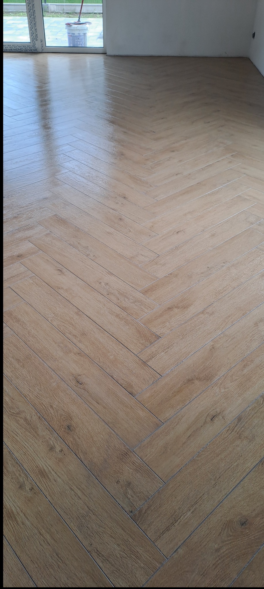 Parkiet ułożony w jodełkę francuską, z widoczną fakturą drewna, w jasnym pomieszczeniu z oknem. Na oknie stoi wiadro z mopem.