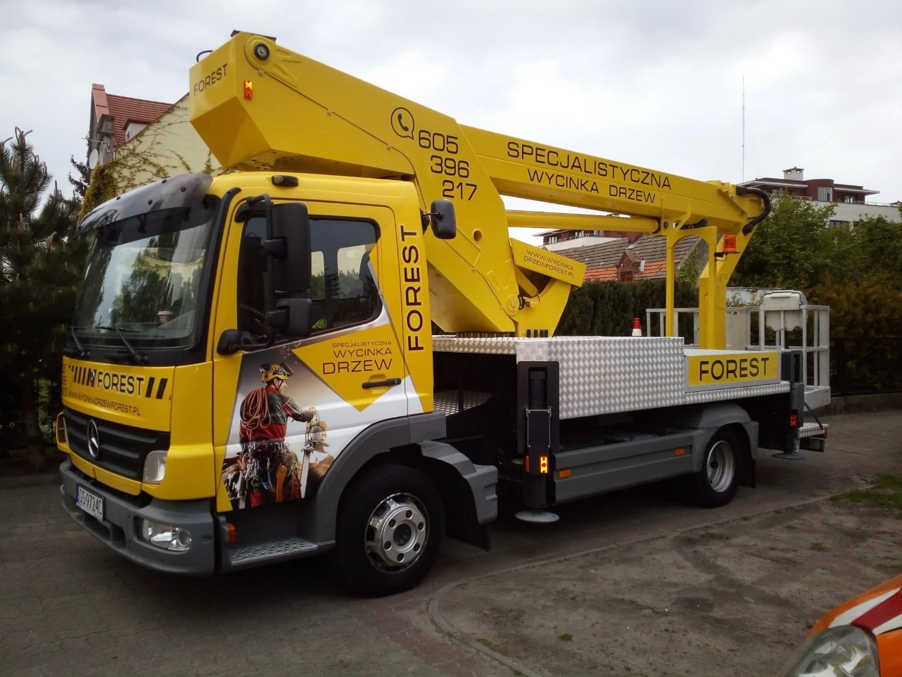 Żółty samochód z podnośnikiem koszowym, z logo firmy FOREST, specjalizującej się w wycince drzew, zaparkowany na tle zabudowań. Widoczne numery kontaktowe.