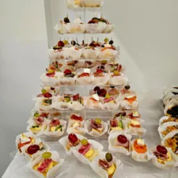 Piramida z apetycznych kanapek na przezroczystych półkach, każda w papierowej foremce, ozdobiona oliwkami, winogronami i wędlinami. Elegancka prezentacja przekąsek.