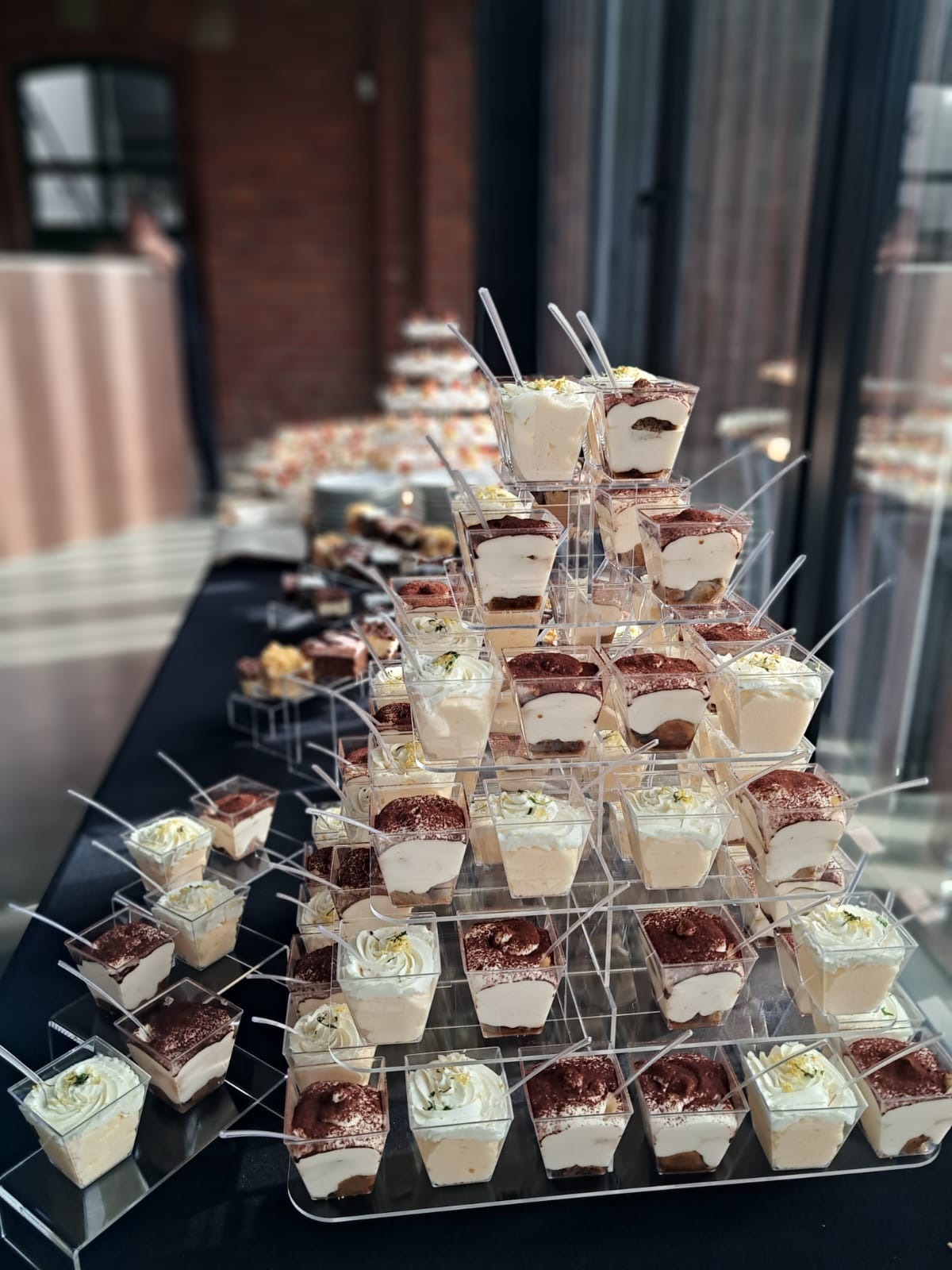 Piramide z deserów w plastikowych pojemnikach, tiramisu i desery z kremem, na czarnym stole, w tle inne słodkości i ceglana ściana. Elegancki catering.