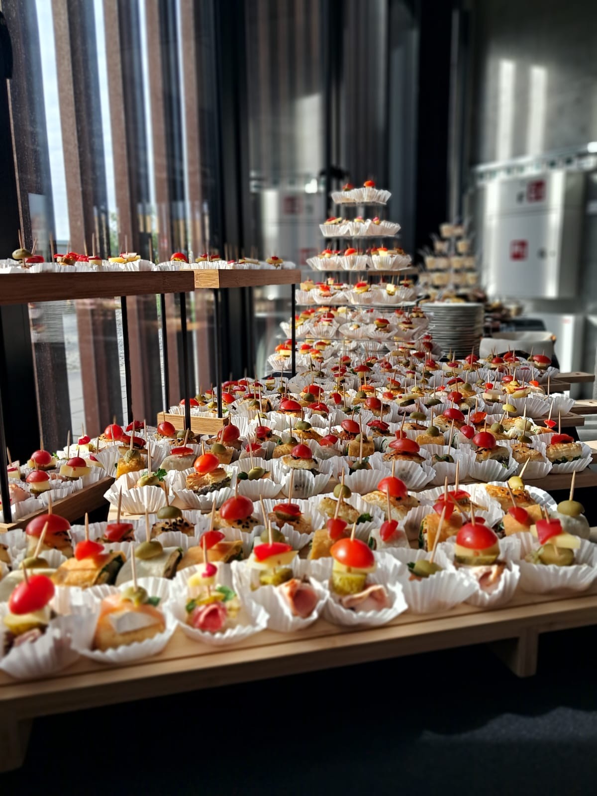 Bogato zastawiony stół z przekąskami cateringowymi, na drewnianych paterach i stojakach. W tle duże okno i stalowe zbiorniki.