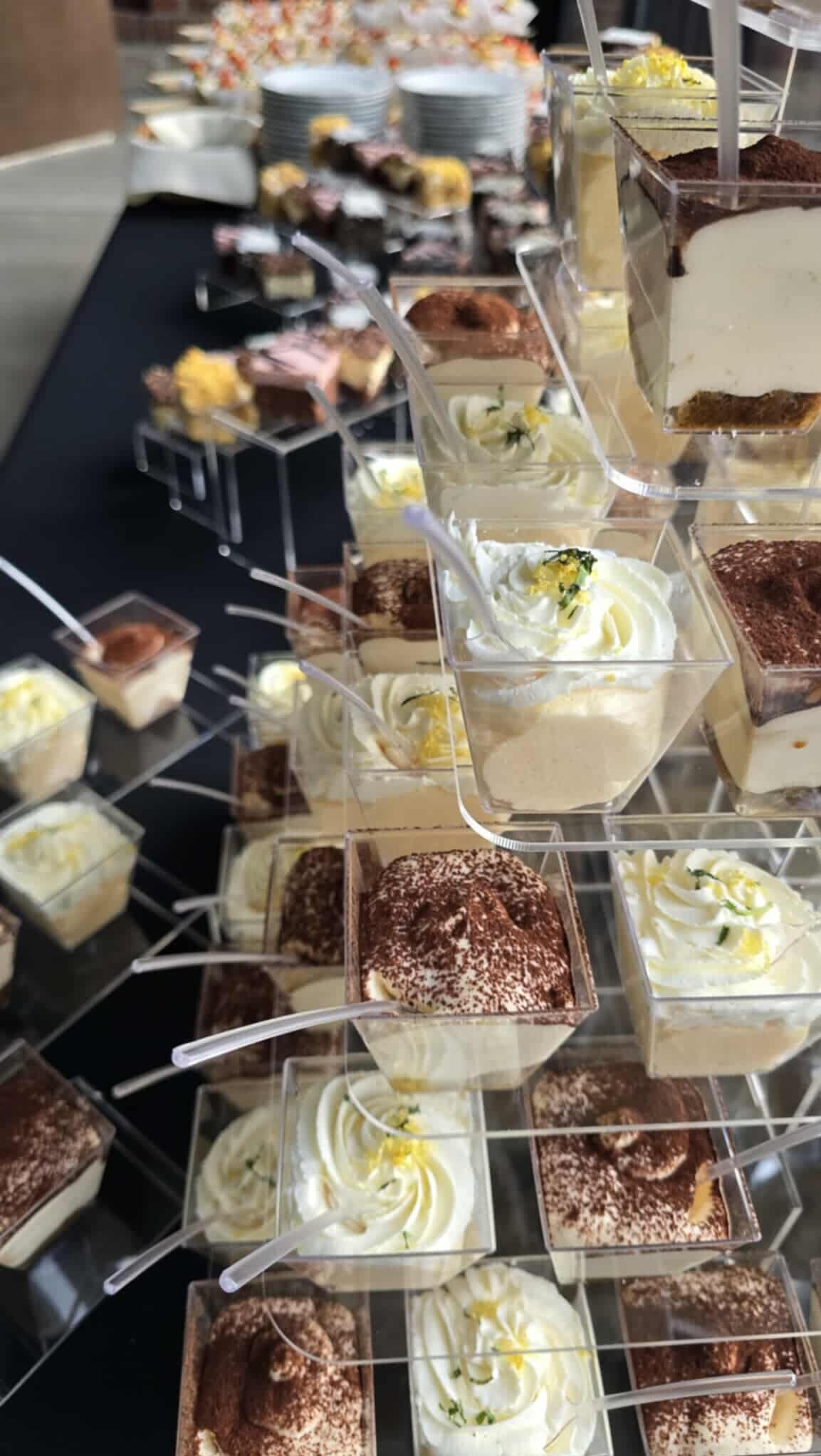 Deserki w przezroczystych pojemnikach, tiramisu i kremy na czarnym stole, gotowe na event. Dekoracyjne, słodkie przekąski na przyjęcie.